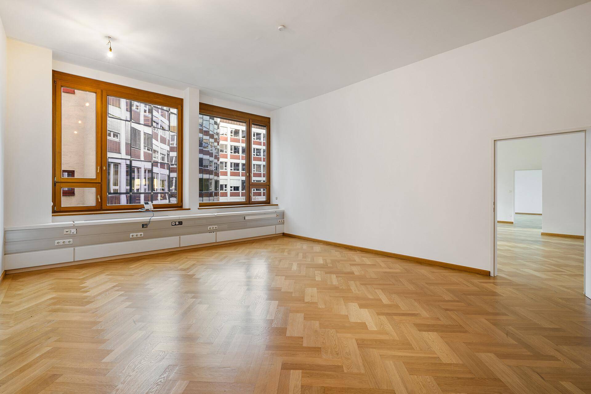 Heller Büroraum mit Fischgrätparkett, weißen Wänden und großen Fenstern mit Stadtblick.