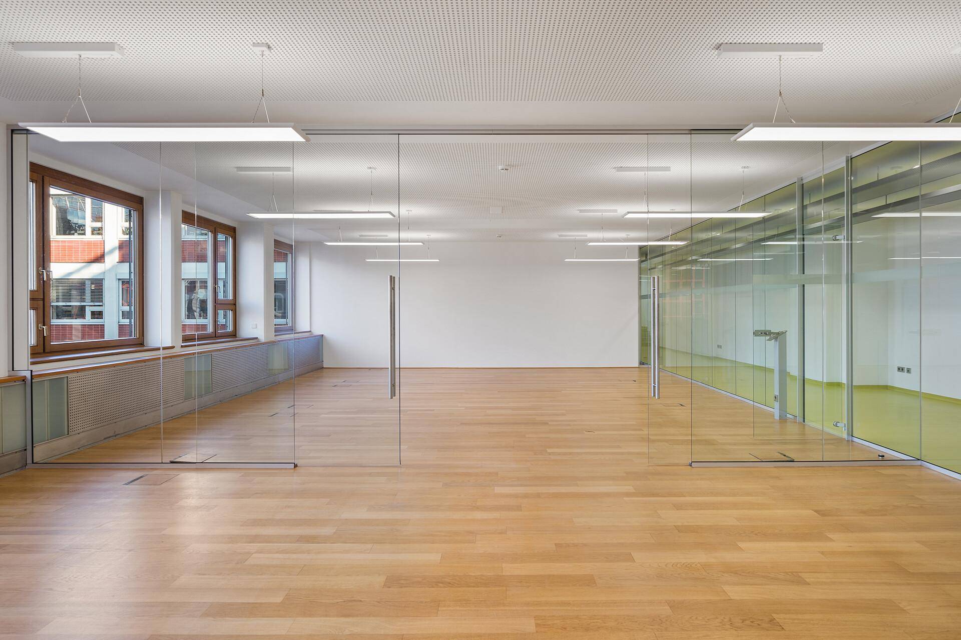 Moderner Bürobereich mit transparenten Glaswänden, Holzboden und viel Tageslicht durch die Fenster.