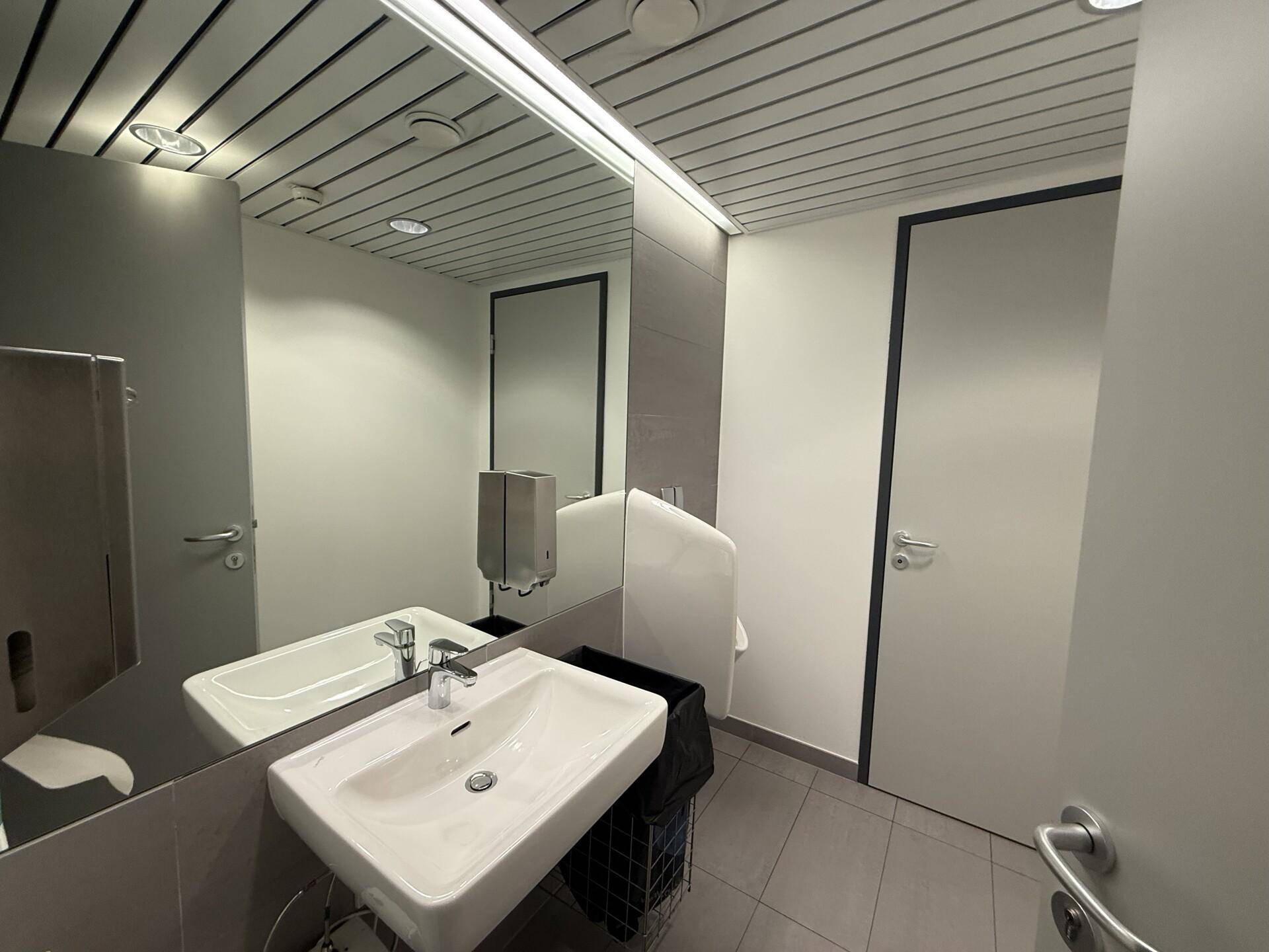 Modernes Badezimmer mit Waschbecken, großem Spiegel und Urinal in neutralen Farben.