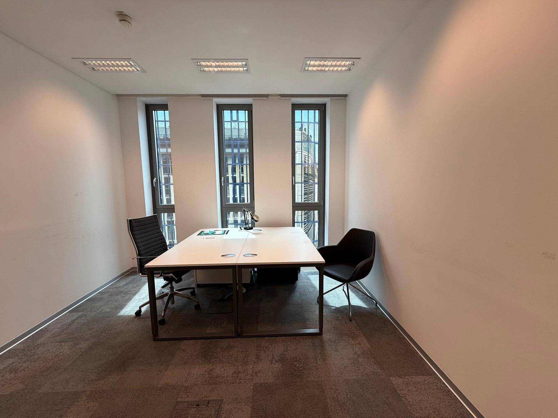 Kleines, helles Büro mit einem Schreibtisch für zwei Personen und Teppichboden.