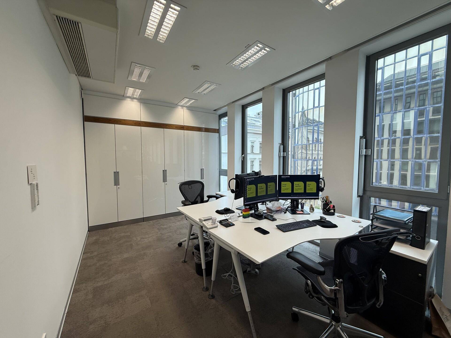 Geräumiges Büro mit mehreren Arbeitsplätzen, integrierten Schränken und großen Fenstern.