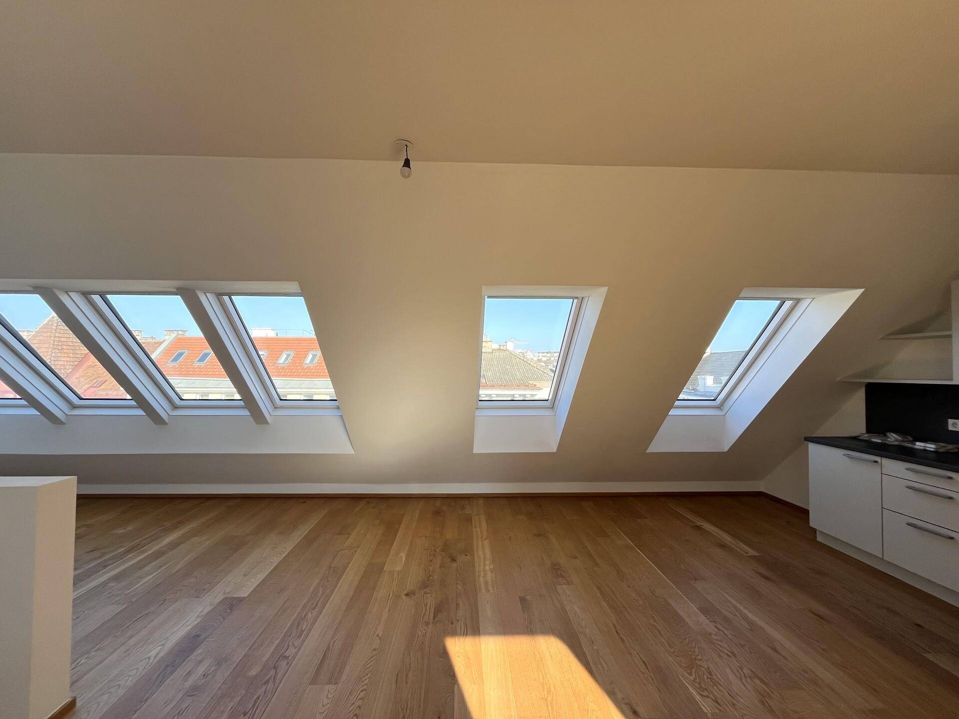 Geräumiger Wohnbereich im Dachgeschoss mit Holzboden, mehreren Dachfenstern und Blick auf die umliegenden Dächer.
