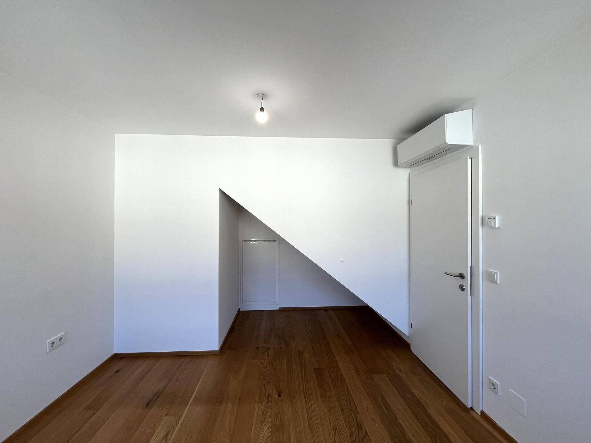 Zimmer mit Holzboden, weißen Wänden, Klimaanlage und einer Tür, die zu einem kleinen Stauraum führt.
