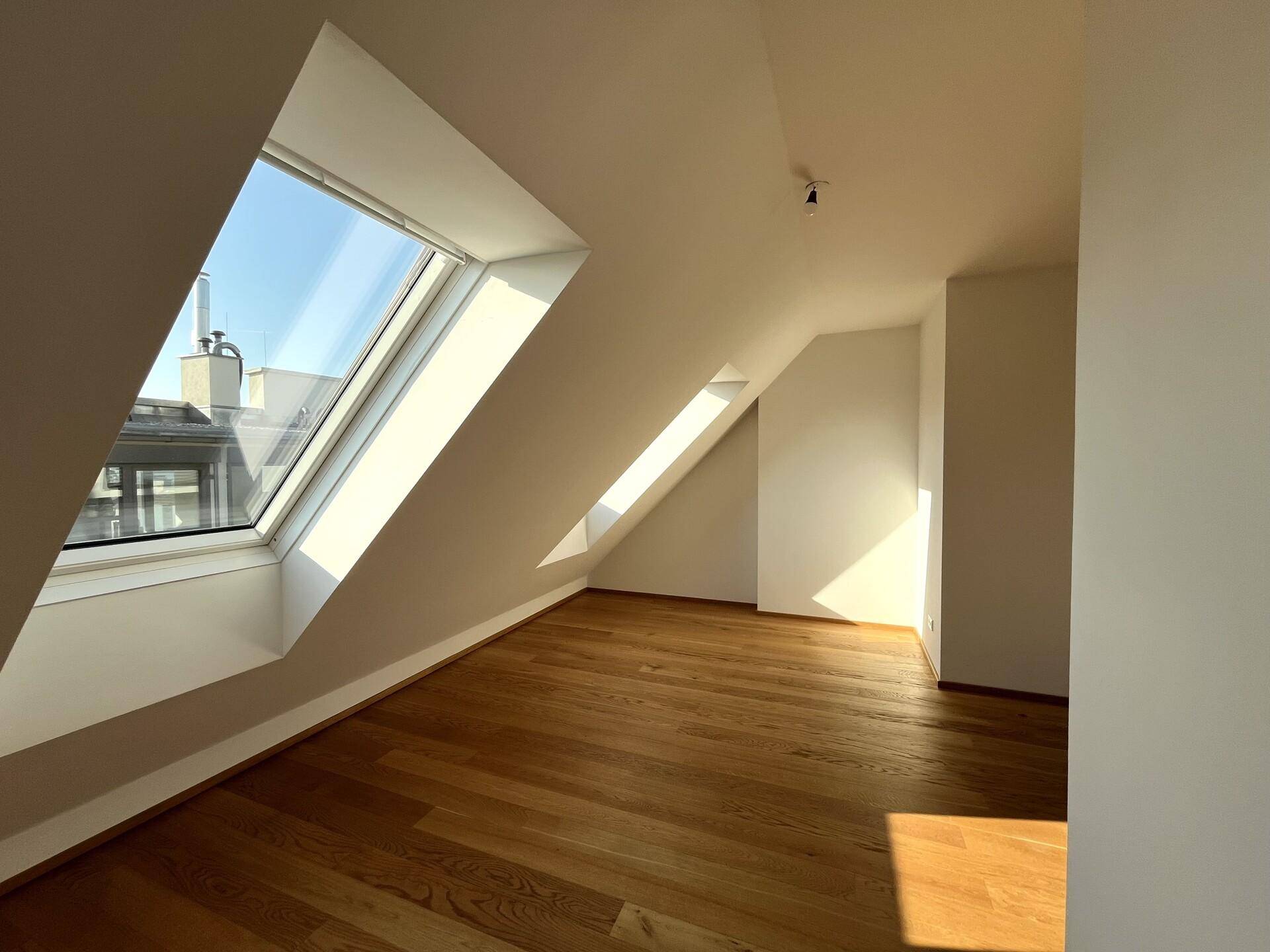 Helles Schlafzimmer im Dachgeschoss mit Holzboden, schrägen Wänden und einem großen Dachfenster für viel Tageslicht.
