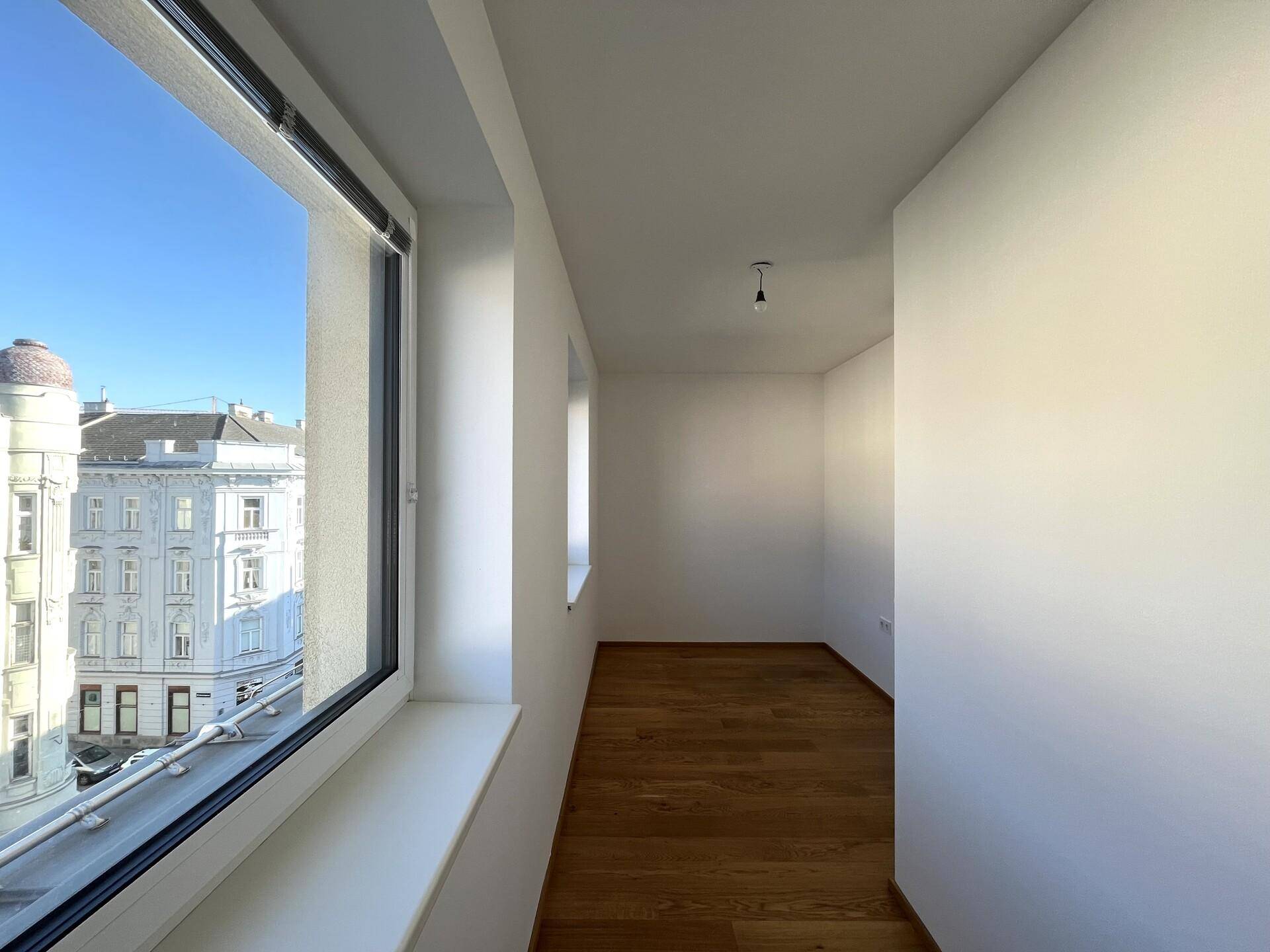 Langer Flur mit Holzboden, weißen Wänden und einem großen Fenster mit Blick auf die Stadt und historische Gebäude.
