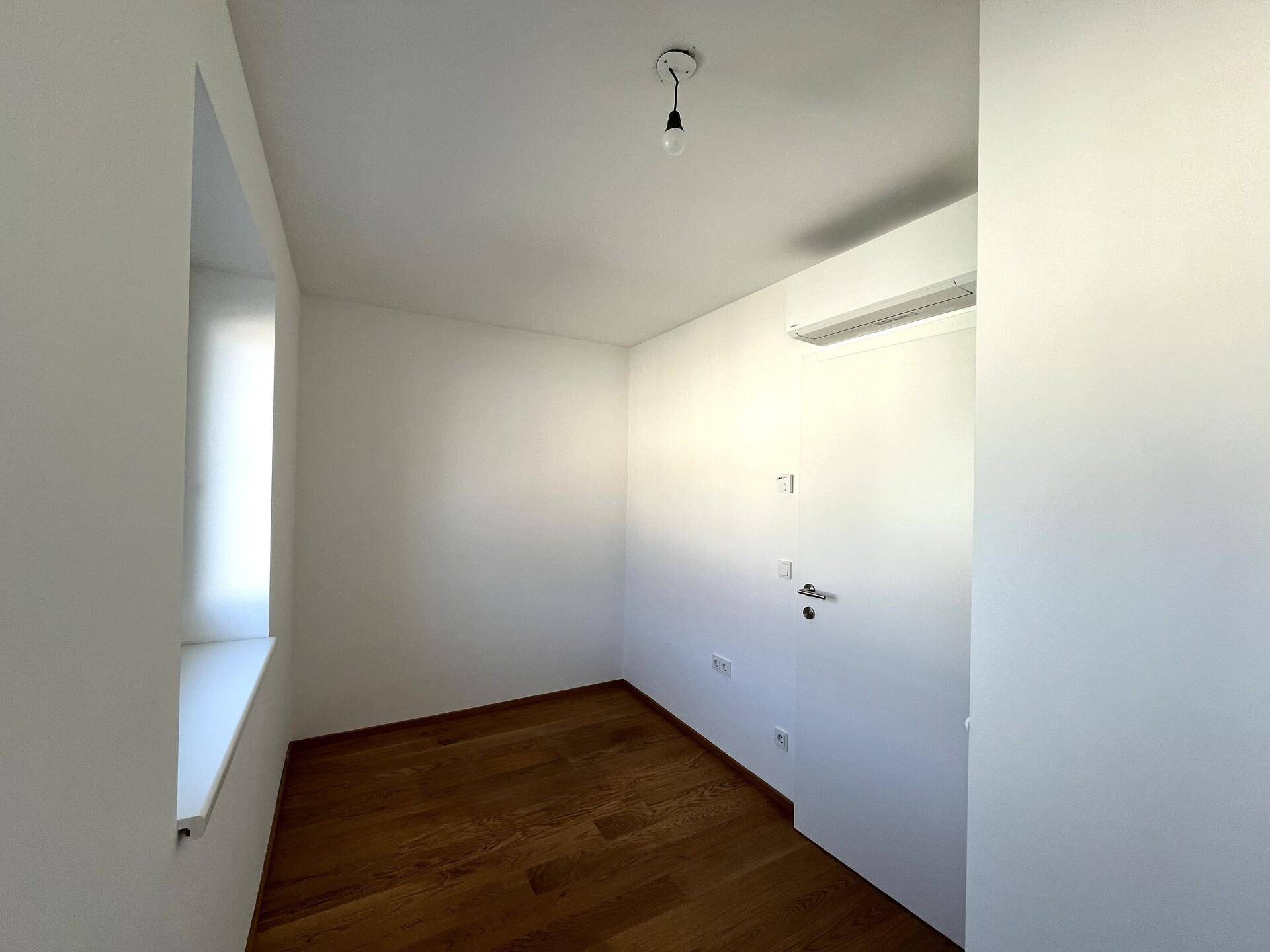 Kleines Zimmer mit Holzboden, weißen Wänden, Klimaanlage und einem Fenster, das Tageslicht hereinlässt.