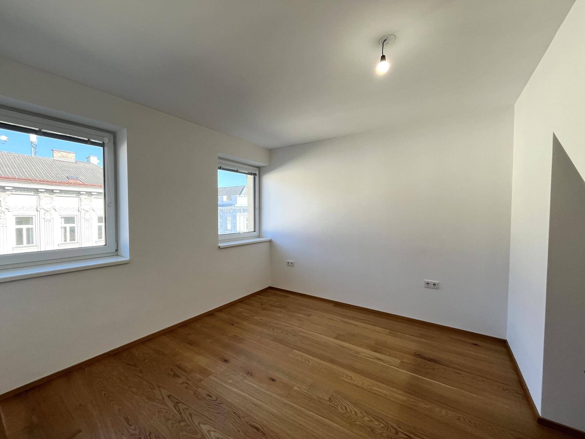 Helles Zimmer mit Holzboden, weißen Wänden und zwei Fenstern, die einen Blick auf die Straße bieten.