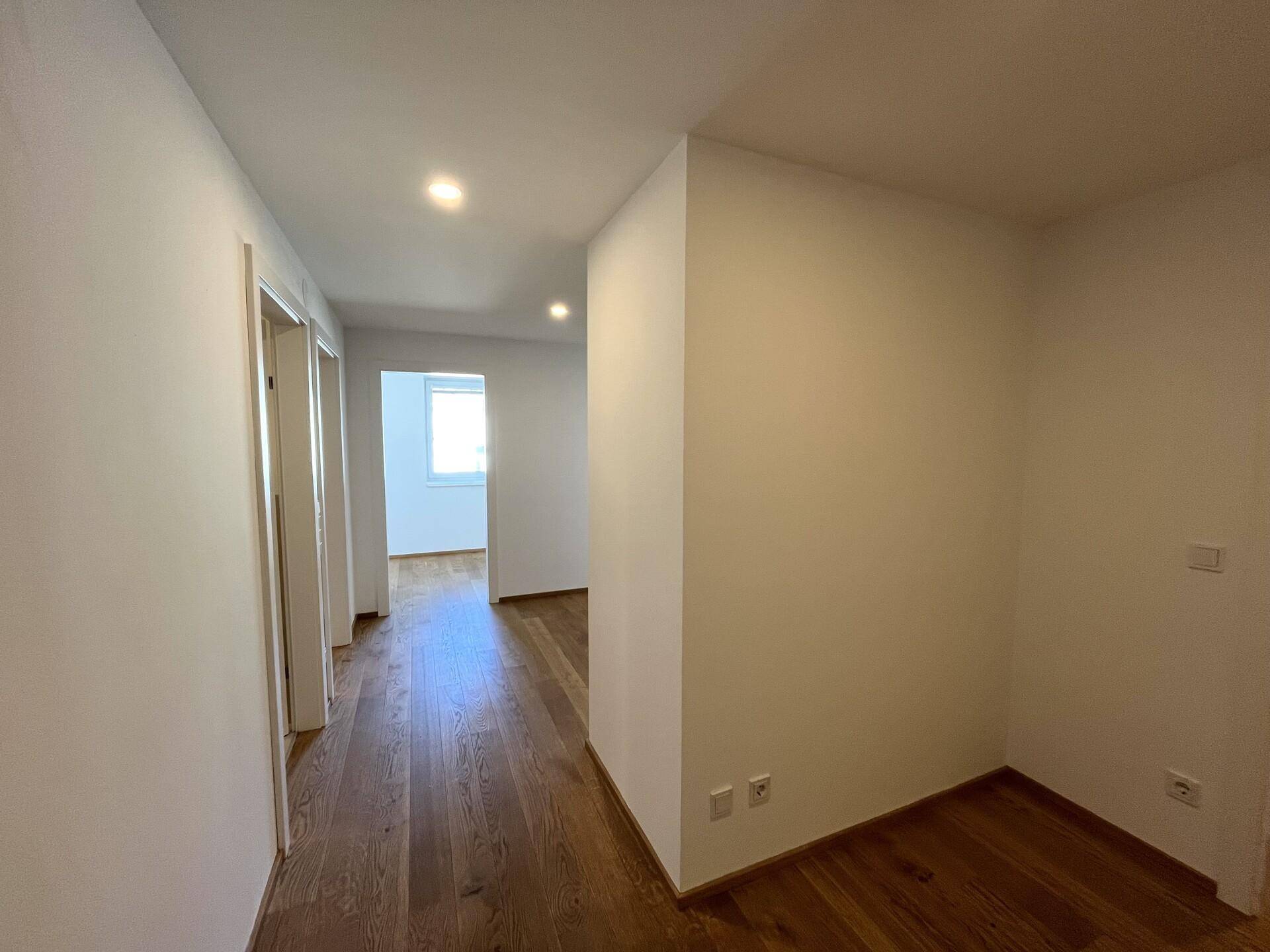 Heller Flur mit Holzboden, weißen Wänden und integrierter Deckenbeleuchtung, der zu weiteren Räumen führt.