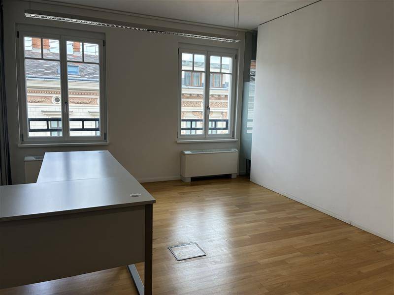Helles, leeres Büro mit Holzfußboden, zwei großen Fenstern und Blick auf die Stadt.