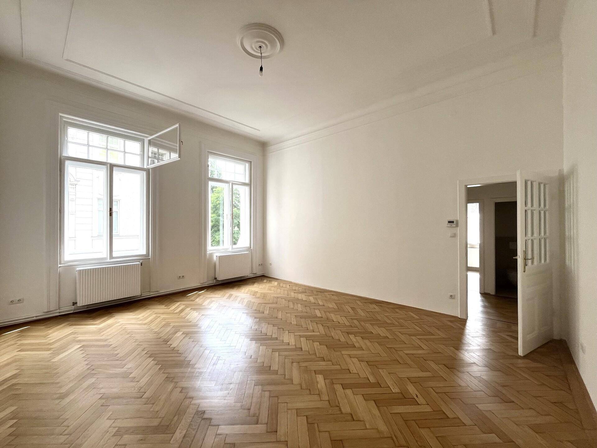 Großes, helles Zimmer mit Fischgrätparkett, hohen Decken und Zugang zu einem weiteren Raum.