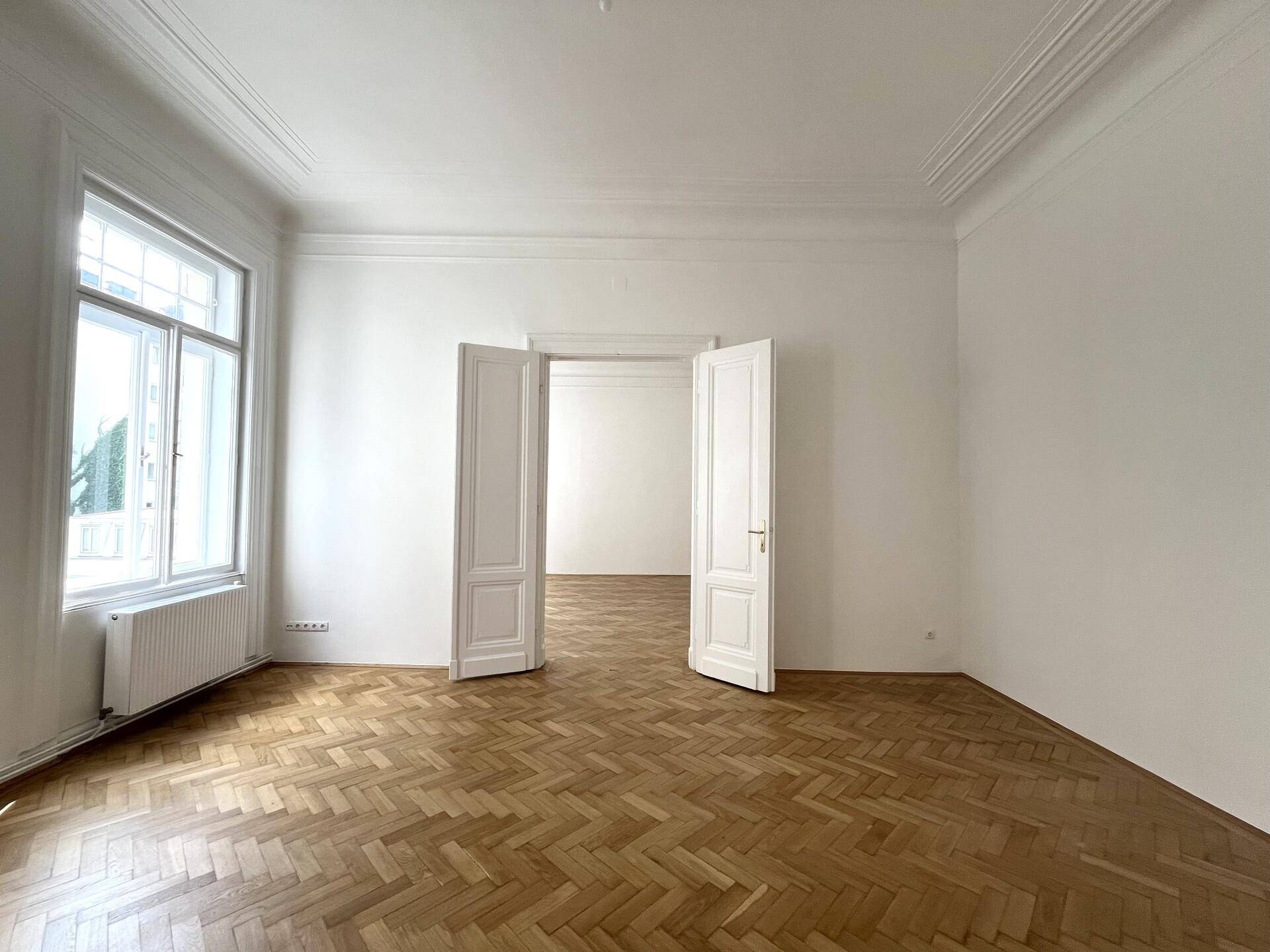 Heller Raum mit Fischgrätparkett, hohen Decken und Flügeltüren zu einem angrenzenden Zimmer.