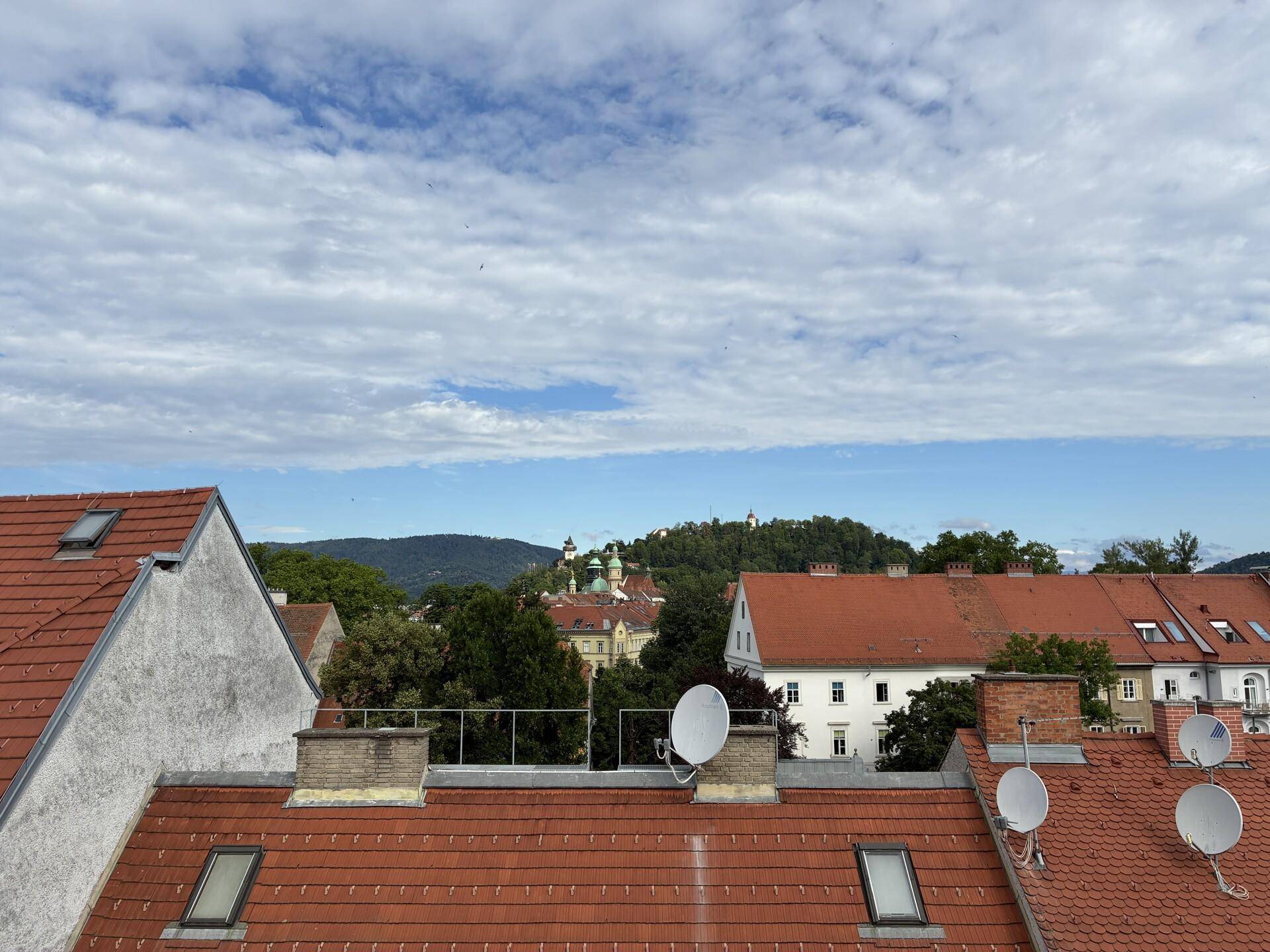 Panoramablick über die Dächer der Stadt mit grünen Hügeln und historischen Gebäuden im Hintergrund.