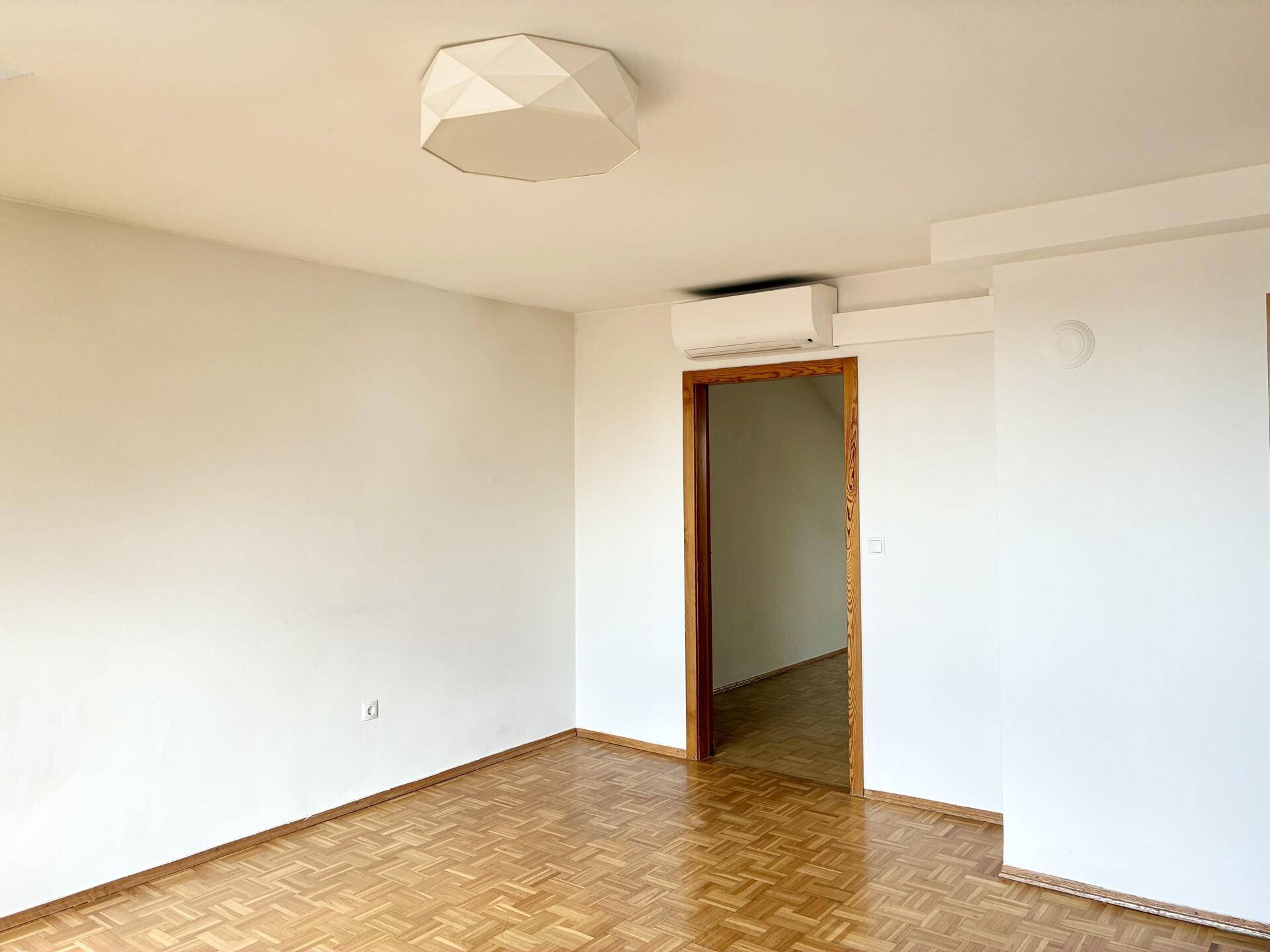 Geräumiges Zimmer mit Parkettboden, weiß gestrichenen Wänden und Klimaanlage.