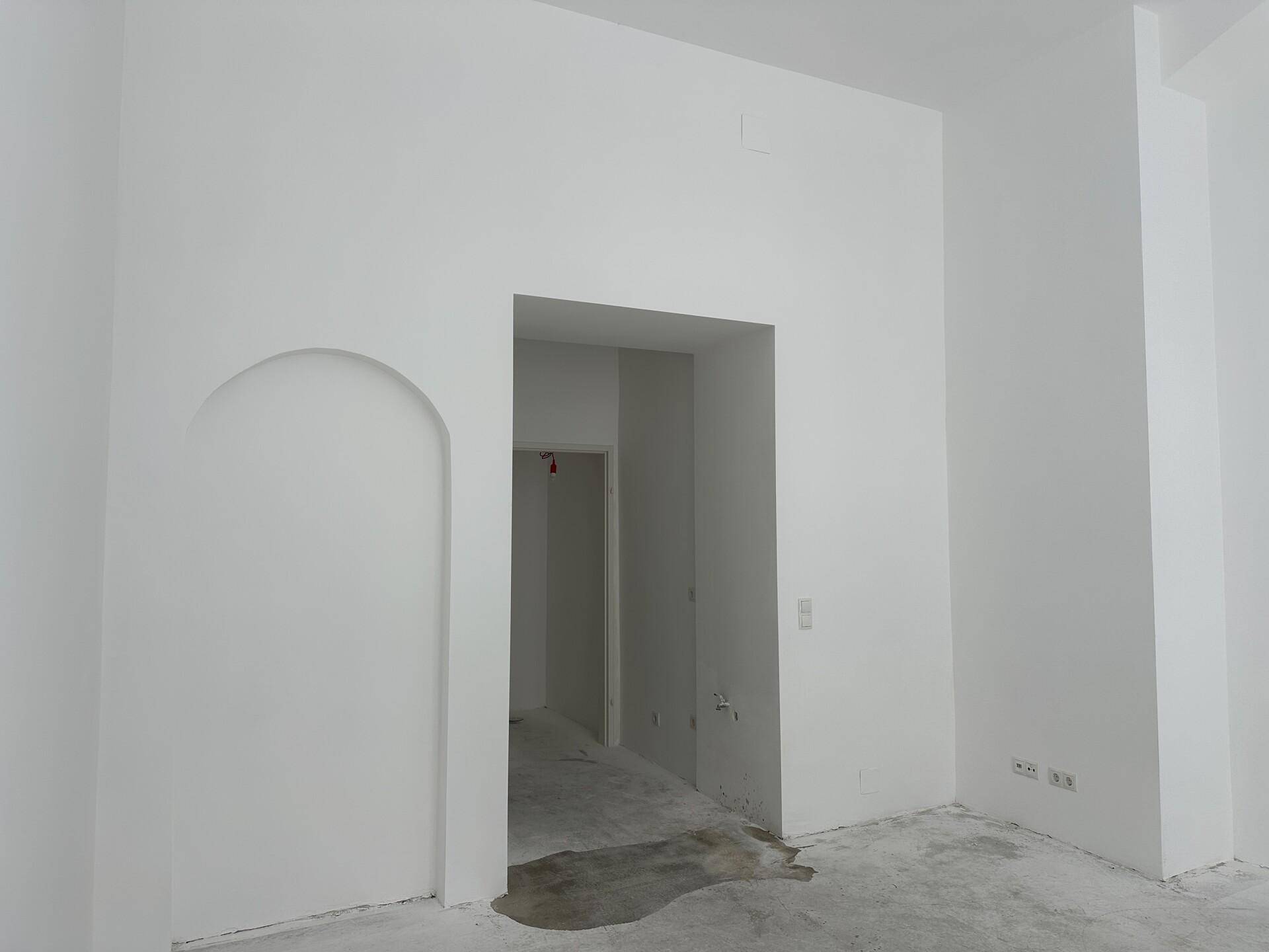 Unmöblierter Raum mit weiß gestrichenen Wänden, Betonboden und einem Durchgang zu weiteren Bereichen.