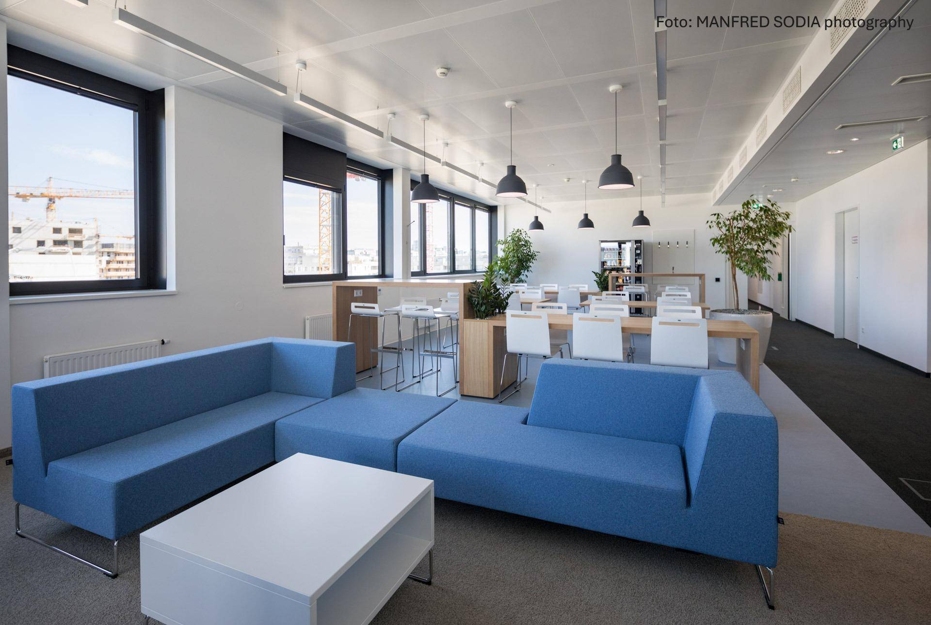 Entspannter Loungebereich in einem Büro mit blauen Sofas, Couchtisch und Blick auf die Stadt.