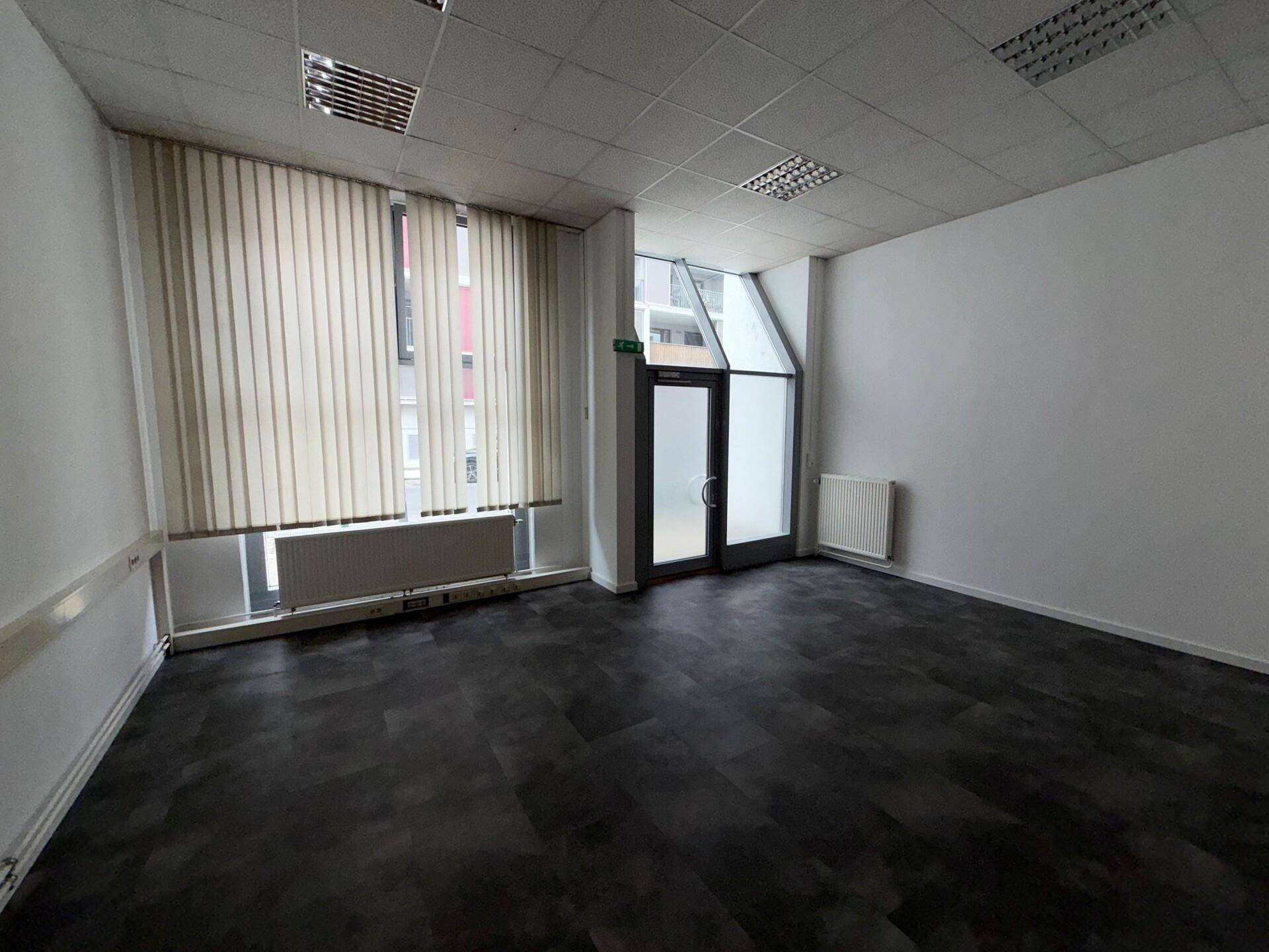 Geräumiger Büroraum mit dunklem Vinylboden, weißen Wänden und großen Fenstern, die viel Tageslicht hereinlassen.