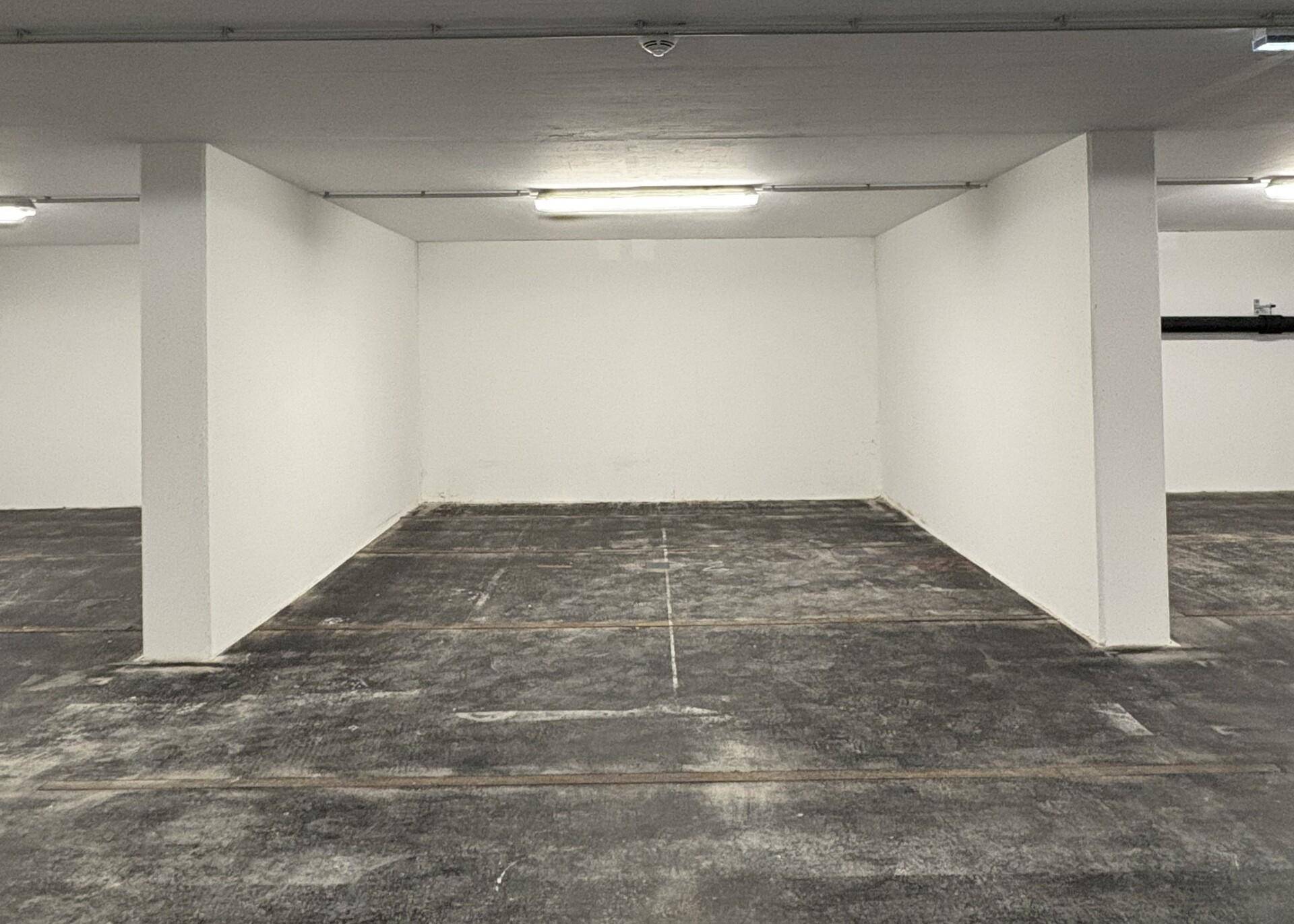 Leerer Parkplatz im Untergeschoss mit dunklem Betonboden, weißen Wänden und sichtbaren Deckeninstallationen.