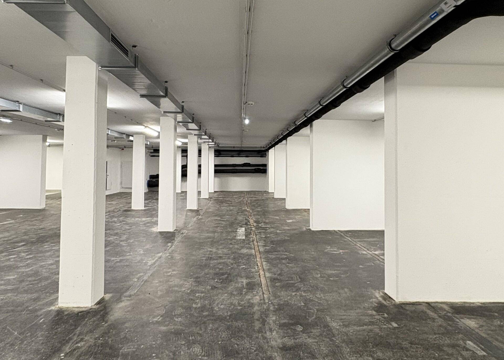 Weitläufige Tiefgarage mit weißen Säulen, dunklem Betonboden und sichtbaren Rohrleitungen an der Decke.