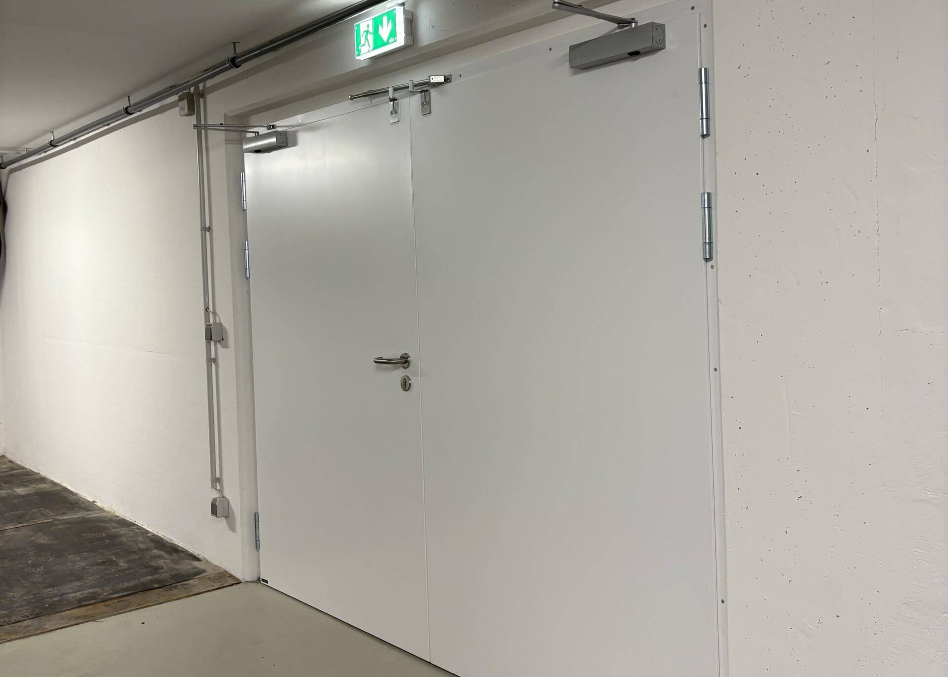 Weiße Doppeltür mit Notausgangsschild in einem Kellerbereich, daneben sichtbare Leitungen an der Wand.