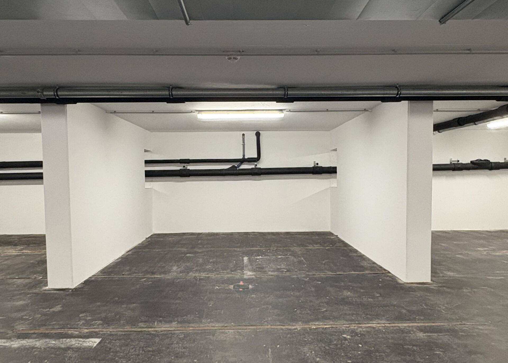 Einzelner Parkplatz oder Lagerbereich mit dunklem Betonboden, weißen Wänden und schwarzen Rohrleitungen an der Decke.