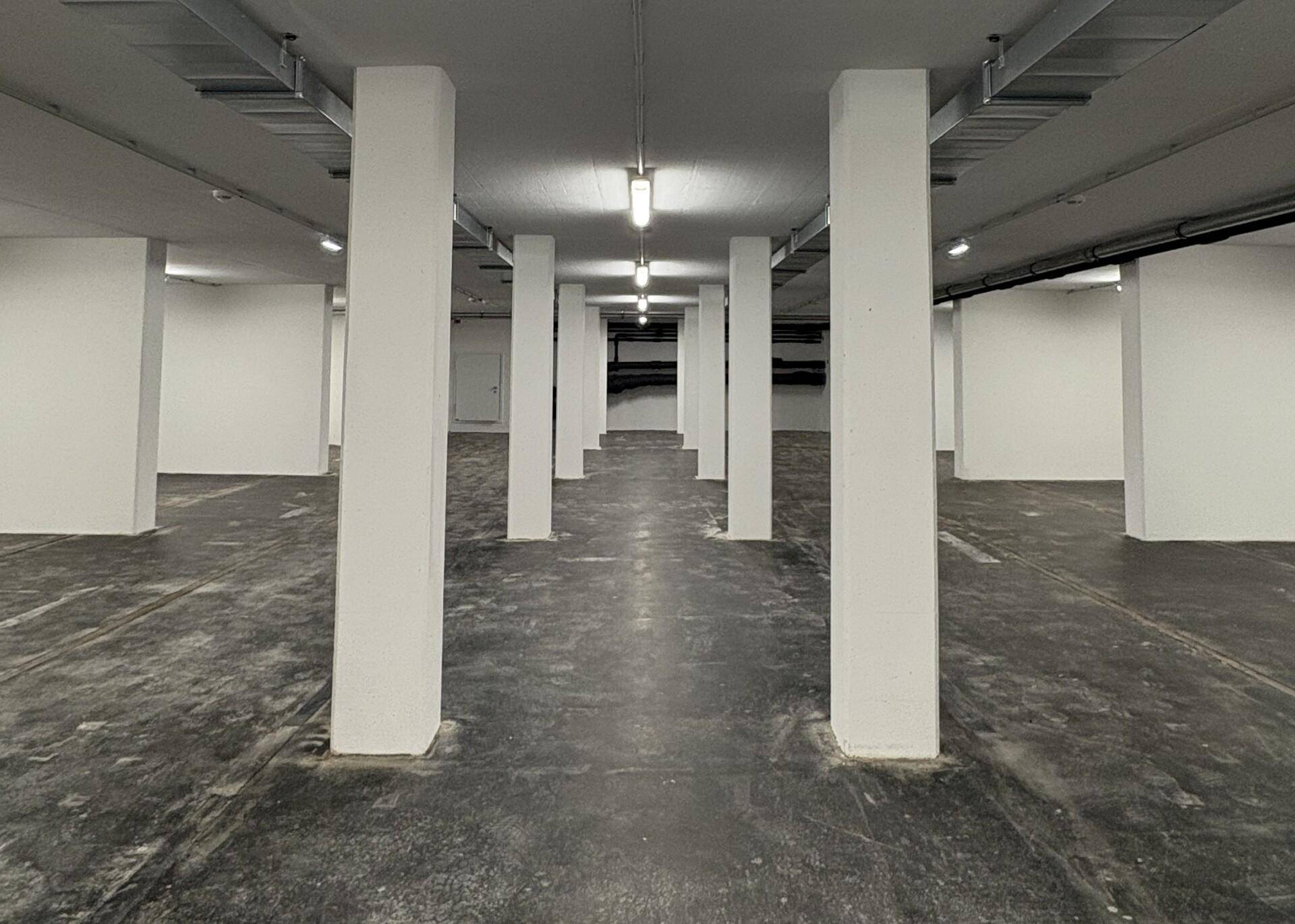 Lange Ansicht einer Tiefgarage mit vielen weißen Säulen, dunklem Betonboden und Reihen von Deckenleuchten.