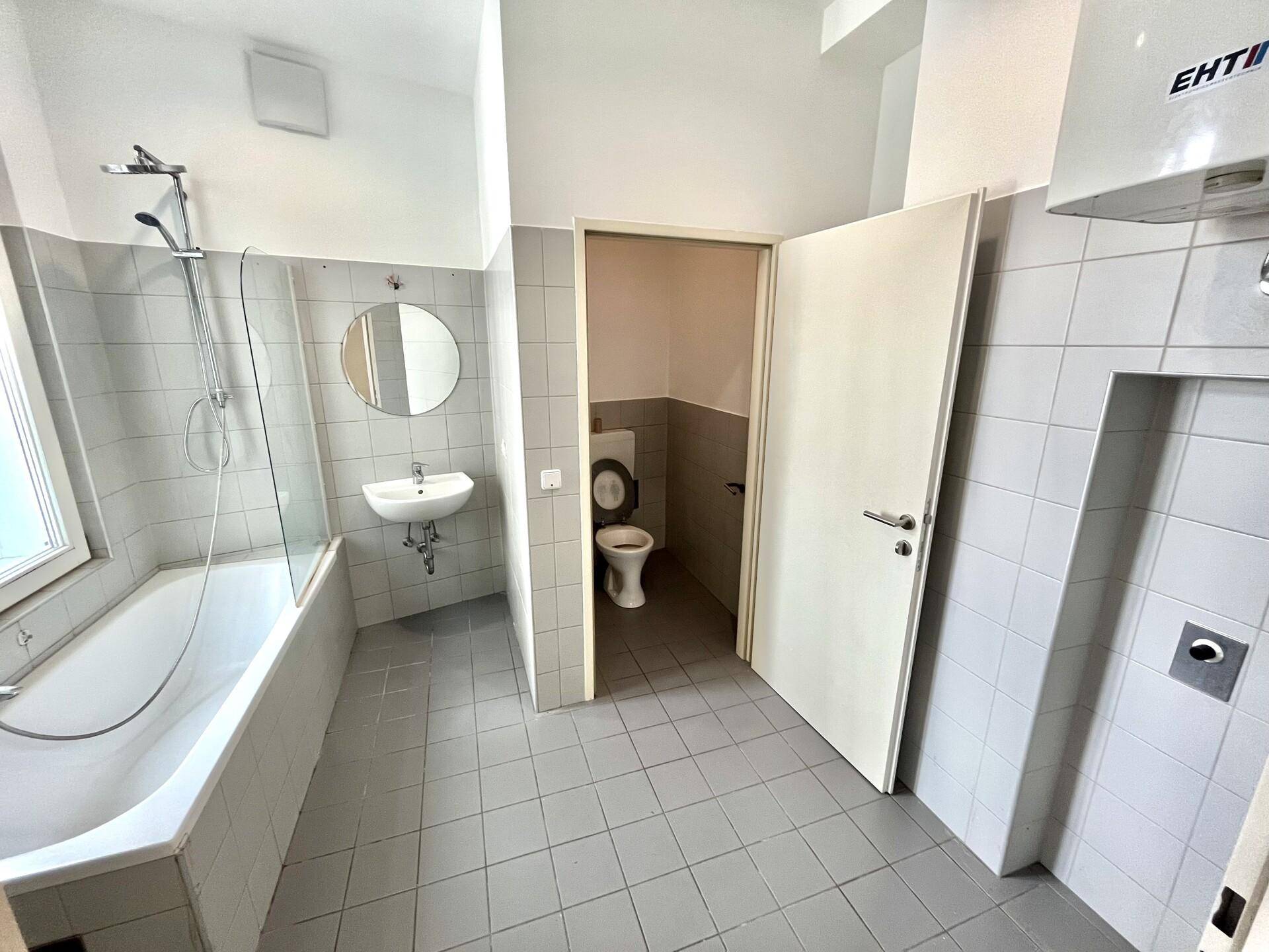 Badezimmer mit Badewanne, Waschbecken und separatem WC, ausgestattet mit hellgrauen Fliesen.