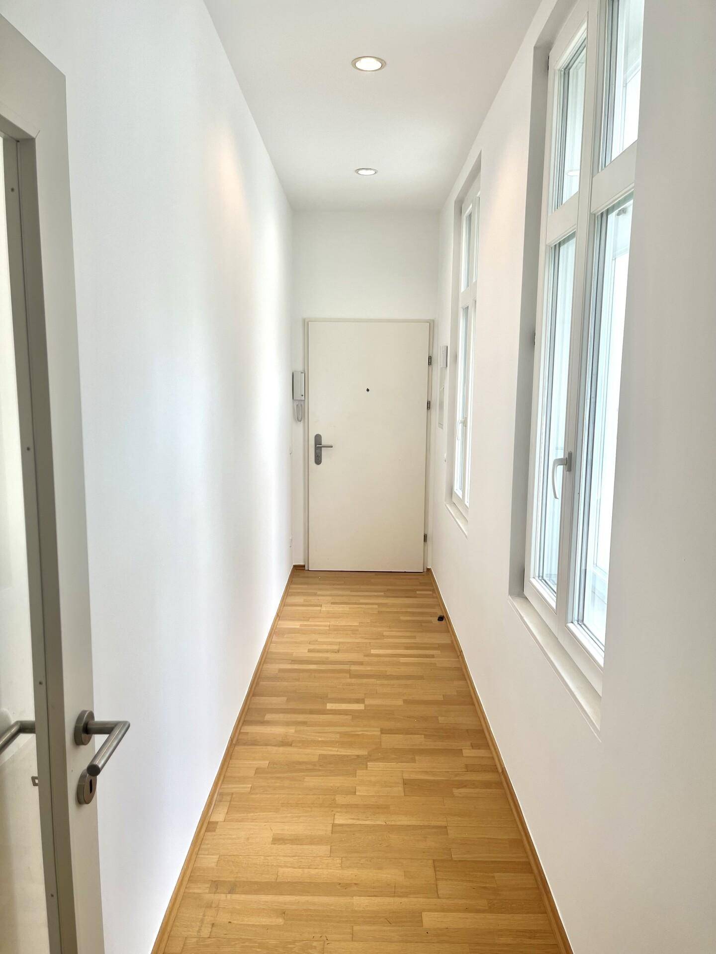 Langer, heller Flur mit Parkettboden, mehreren Fenstern und einer Eingangstür am Ende.