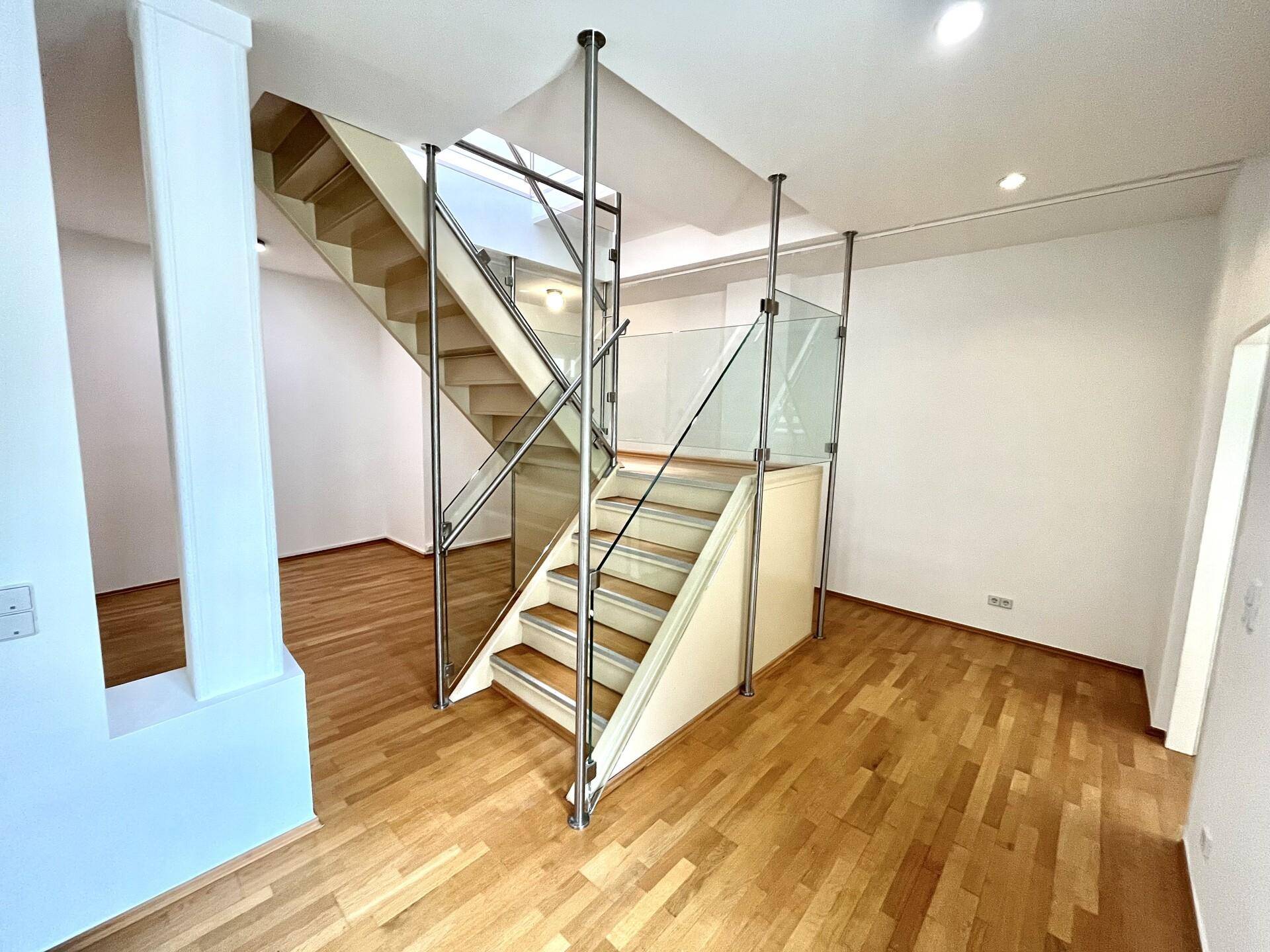 Moderne Holztreppe mit Glasgeländer als zentrales Element in einem hellen Wohnbereich mit Parkettboden.