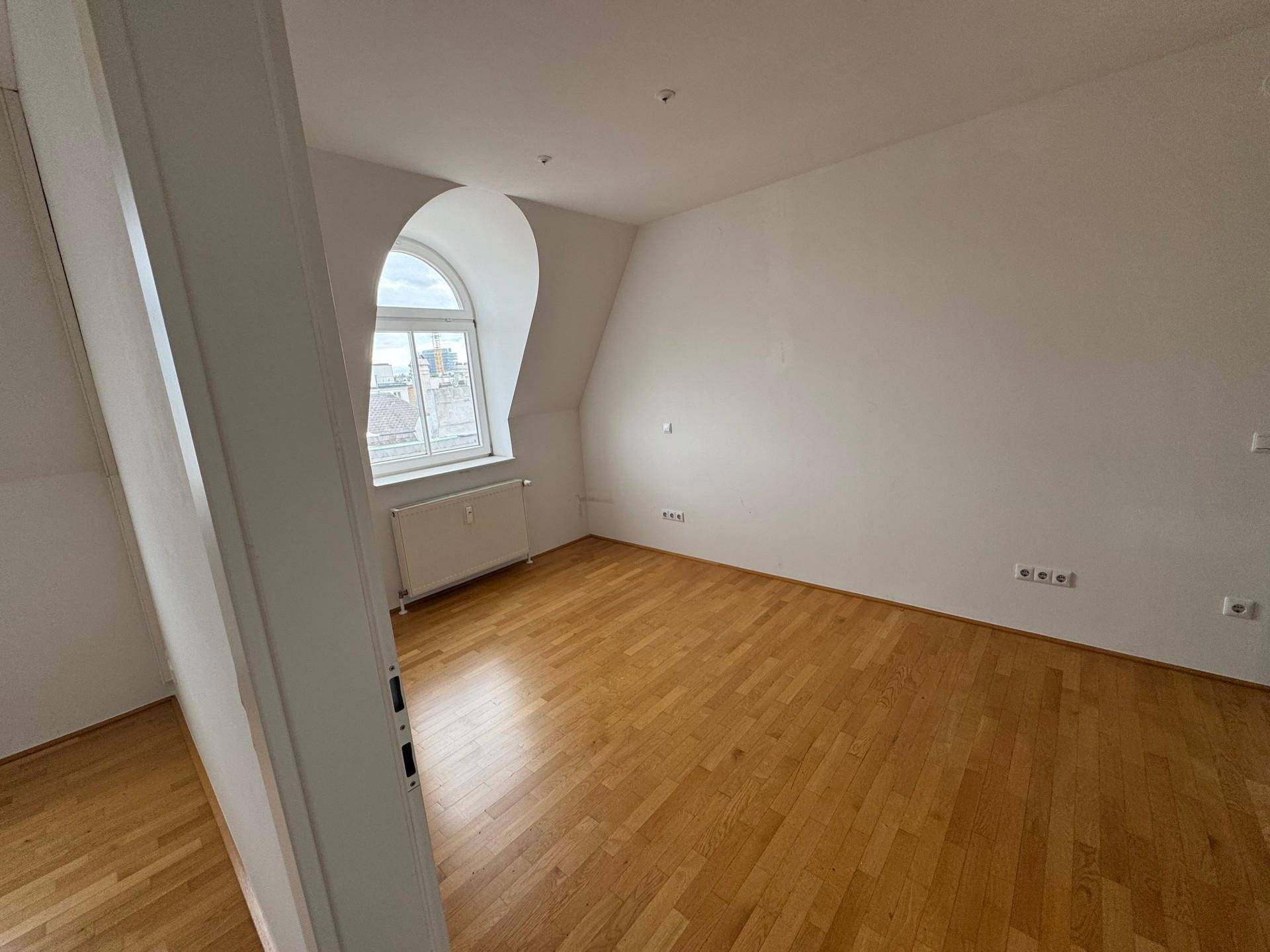 Helles Zimmer mit Holzboden, weiß gestrichenen Wänden und einem markanten Bogenfenster.