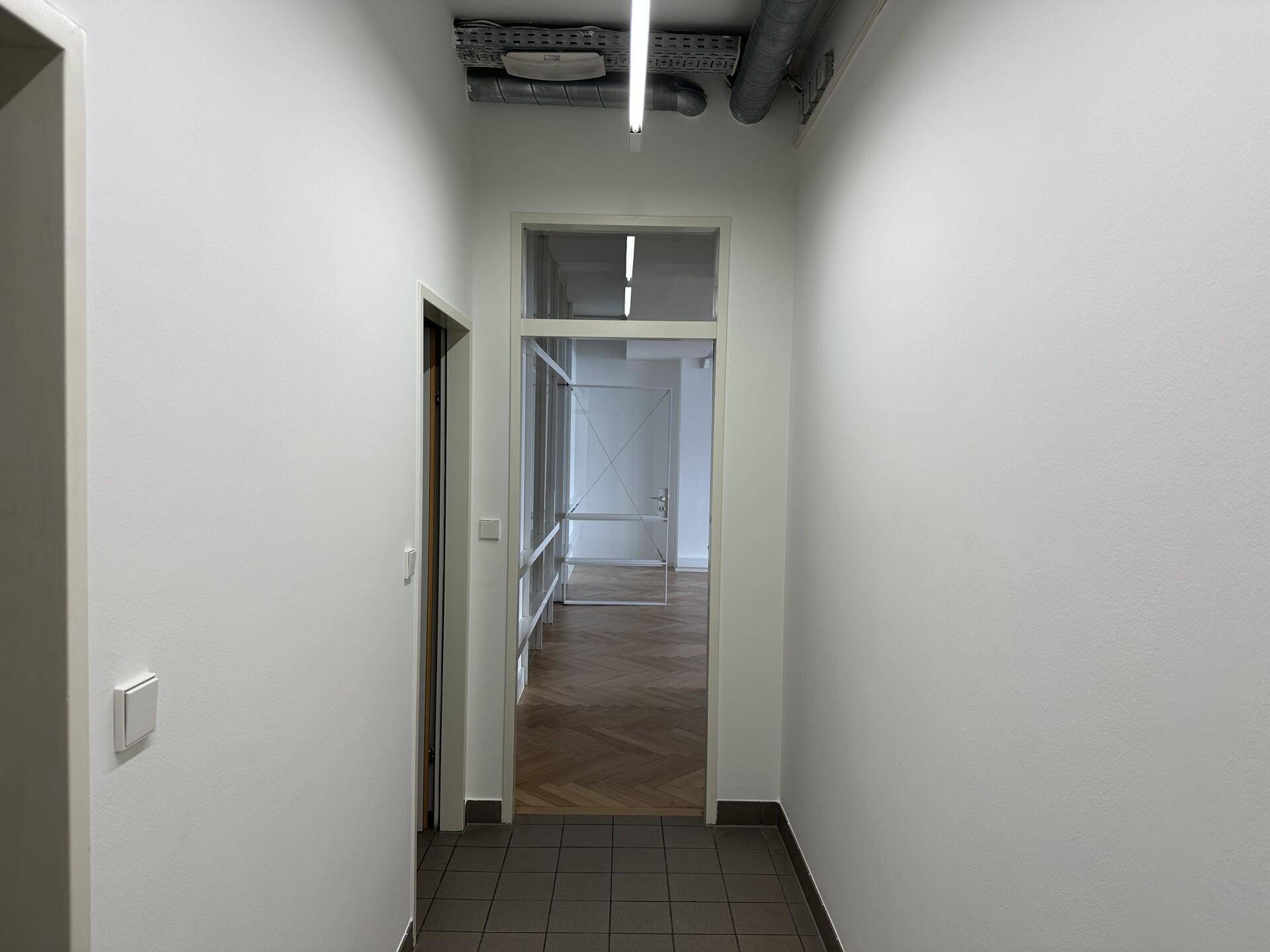 Heller Flur mit Fliesenboden und sichtbaren Installationen an der Decke, führt zu weiteren Räumen.