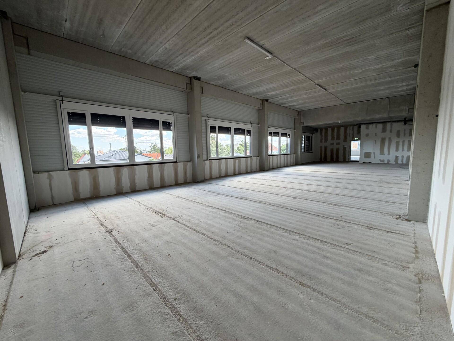 Unfertiger Büroraum mit Betonwänden, Betonboden und mehreren Fenstern mit Rollläden.
