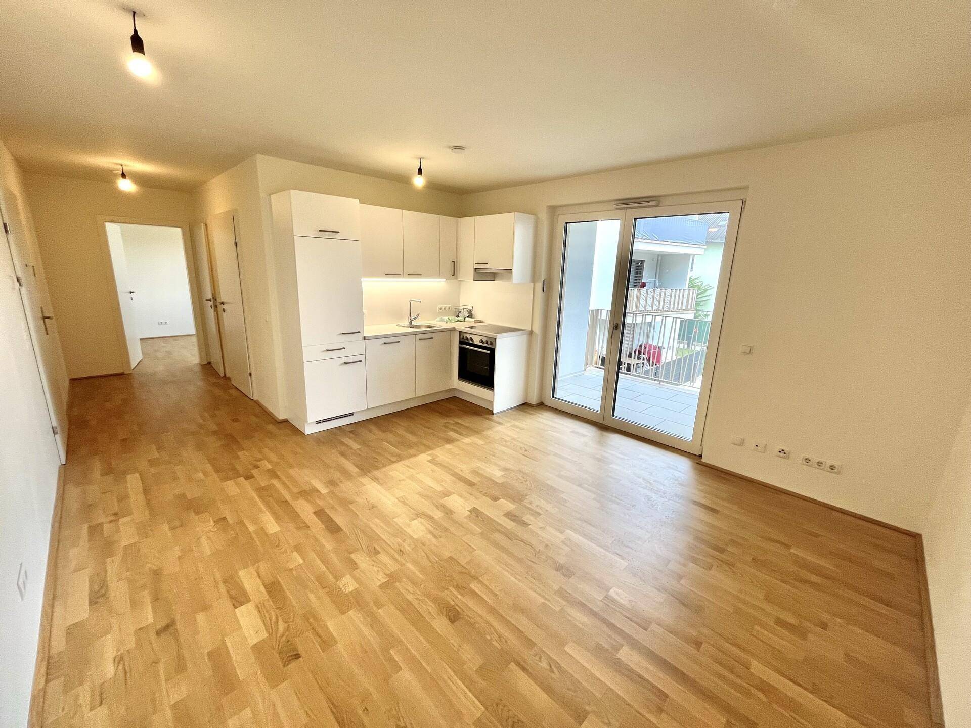 Heller offener Wohnbereich mit integrierter Küchenzeile, Parkettboden und Zugang zum Balkon.