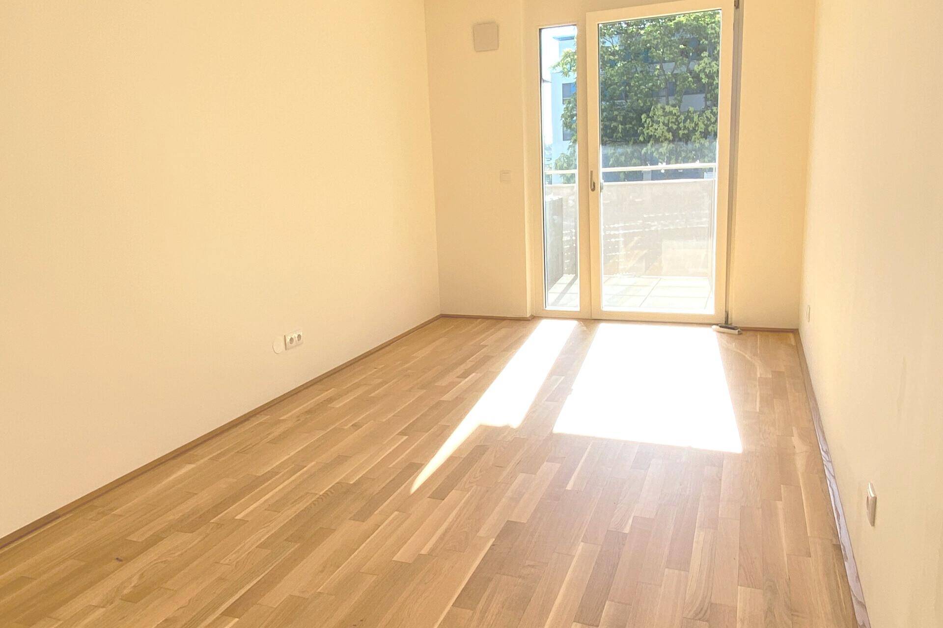Helles Zimmer mit Parkettboden und Balkonzugang, von Sonnenlicht durchflutet.
