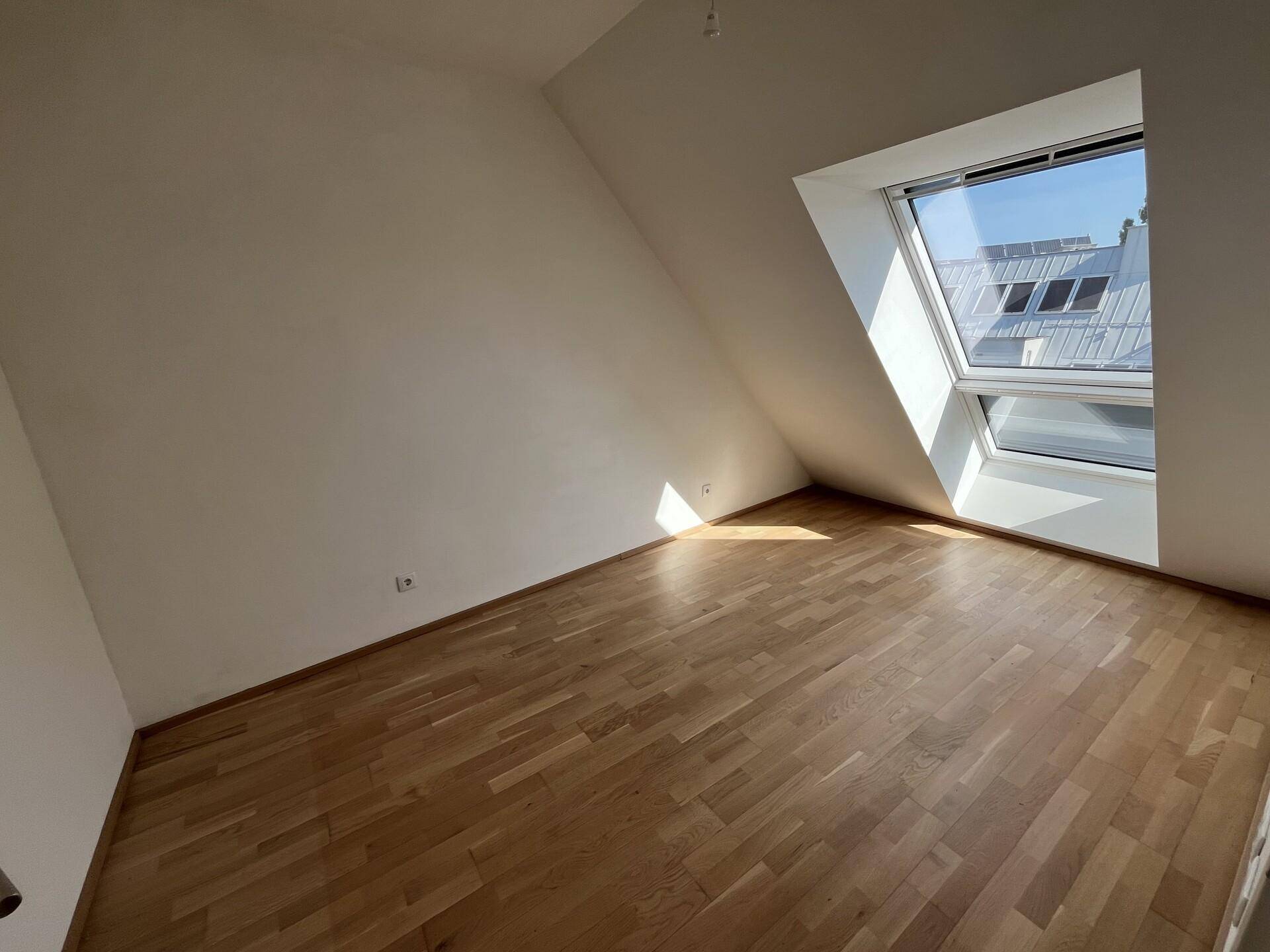 Helles Zimmer im Dachgeschoss mit schräger Decke, großem Dachfenster und Holzparkettboden.