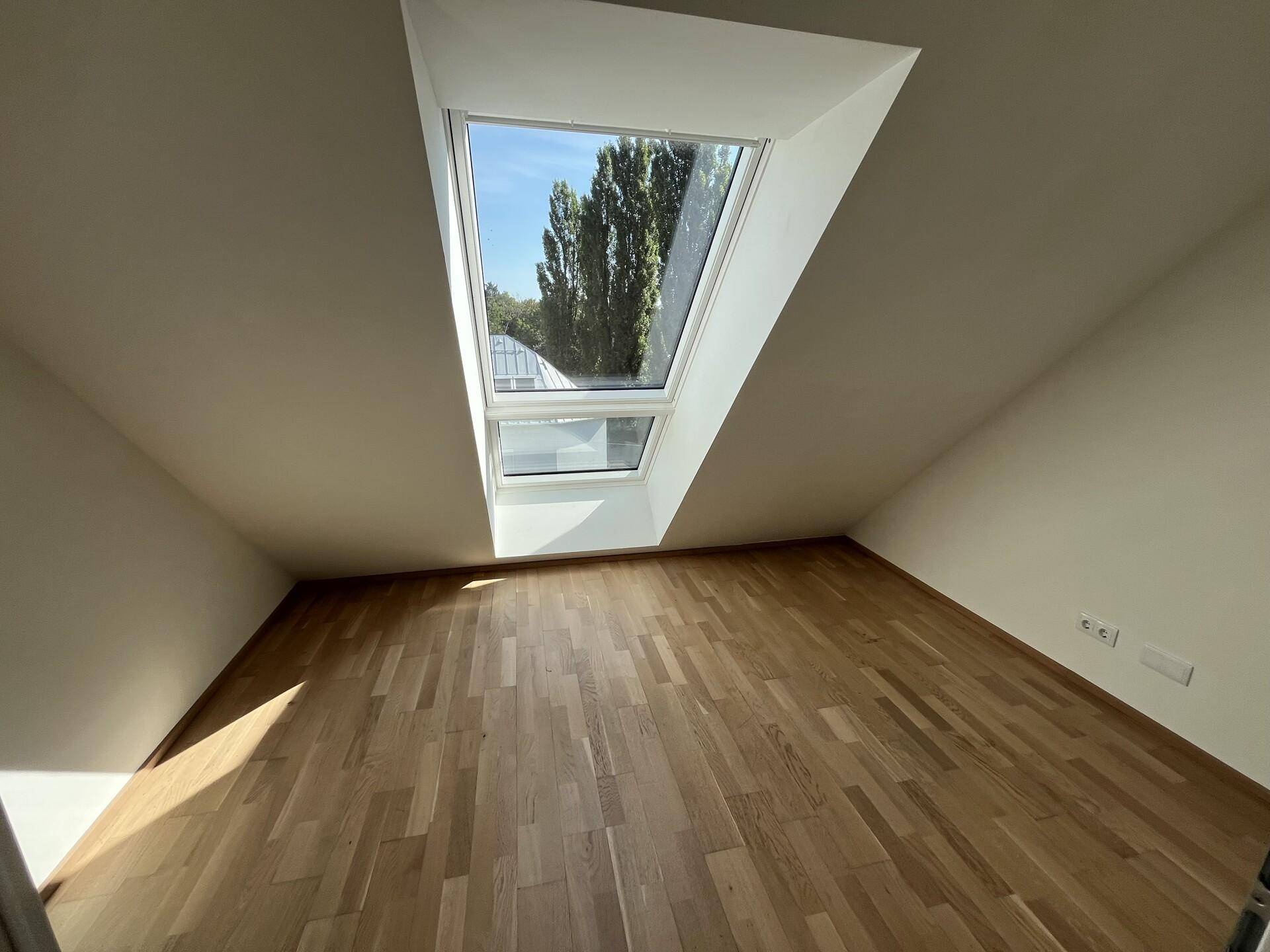 Lichtdurchflutetes Dachzimmer mit schräger Decke, großem Dachfenster und Blick ins Grüne.