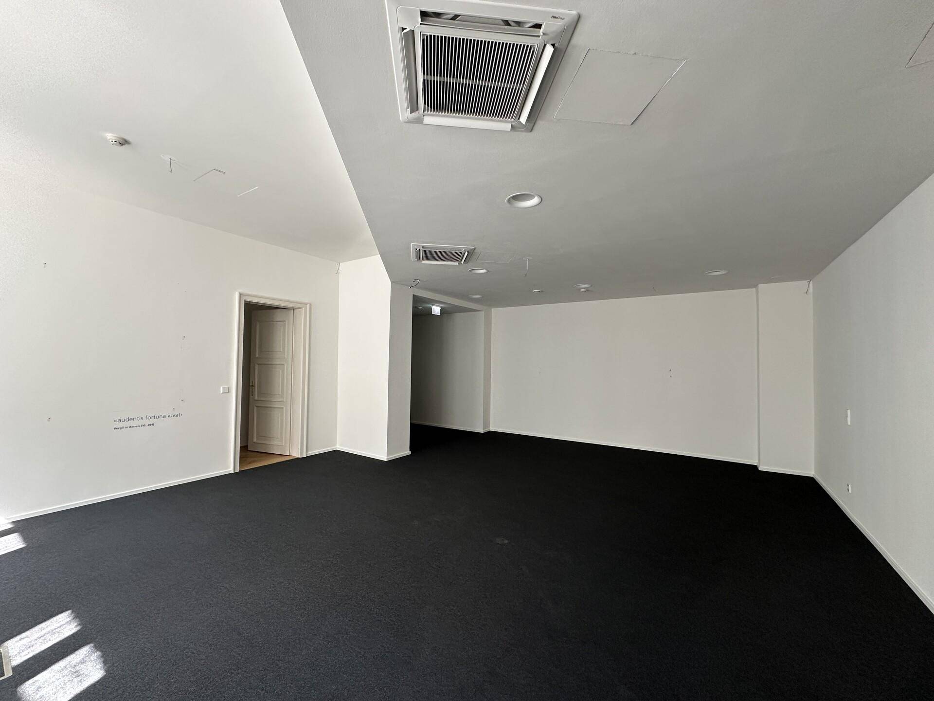 Geräumiger, leerer Büroraum mit dunklem Teppichboden und integrierten Klimaanlagen in der Decke.