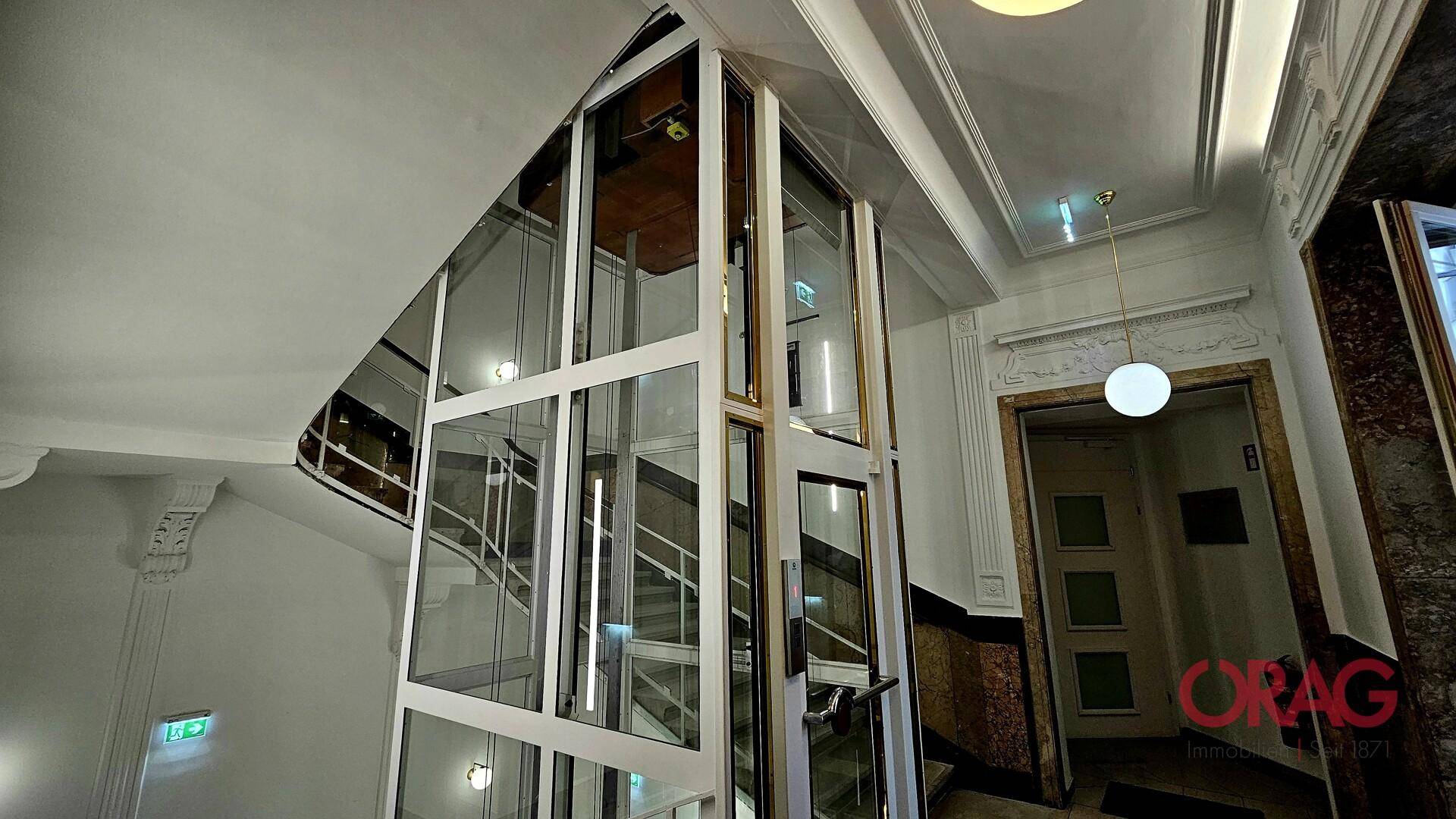 Heller Treppenaufgang mit einem modernen Glasaufzug, der die verschiedenen Etagen des Gebäudes verbindet.