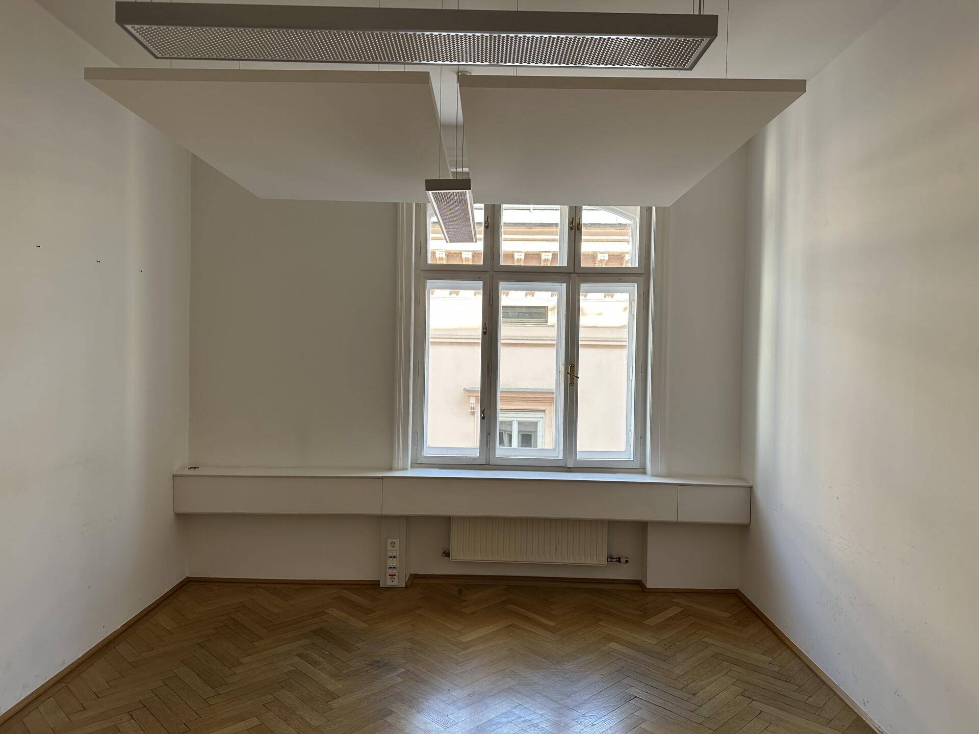 Helles Büro mit Fischgrätparkett, großem Fenster und schwebenden Akustikpaneelen an der Decke.