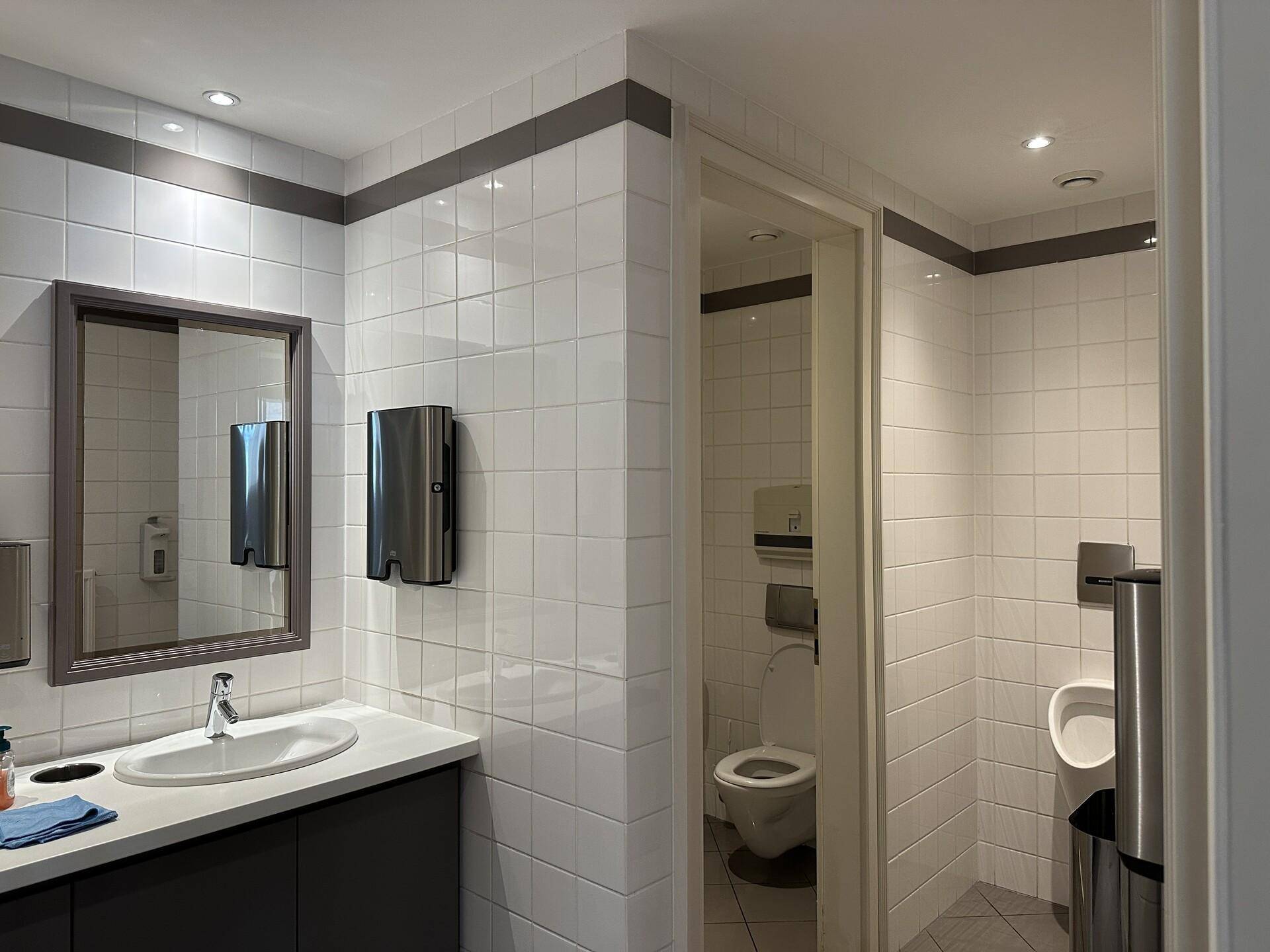 Moderner Waschraum mit weißen Fliesen, einem Waschbecken, Spiegel und separater Toilette mit Urinal.