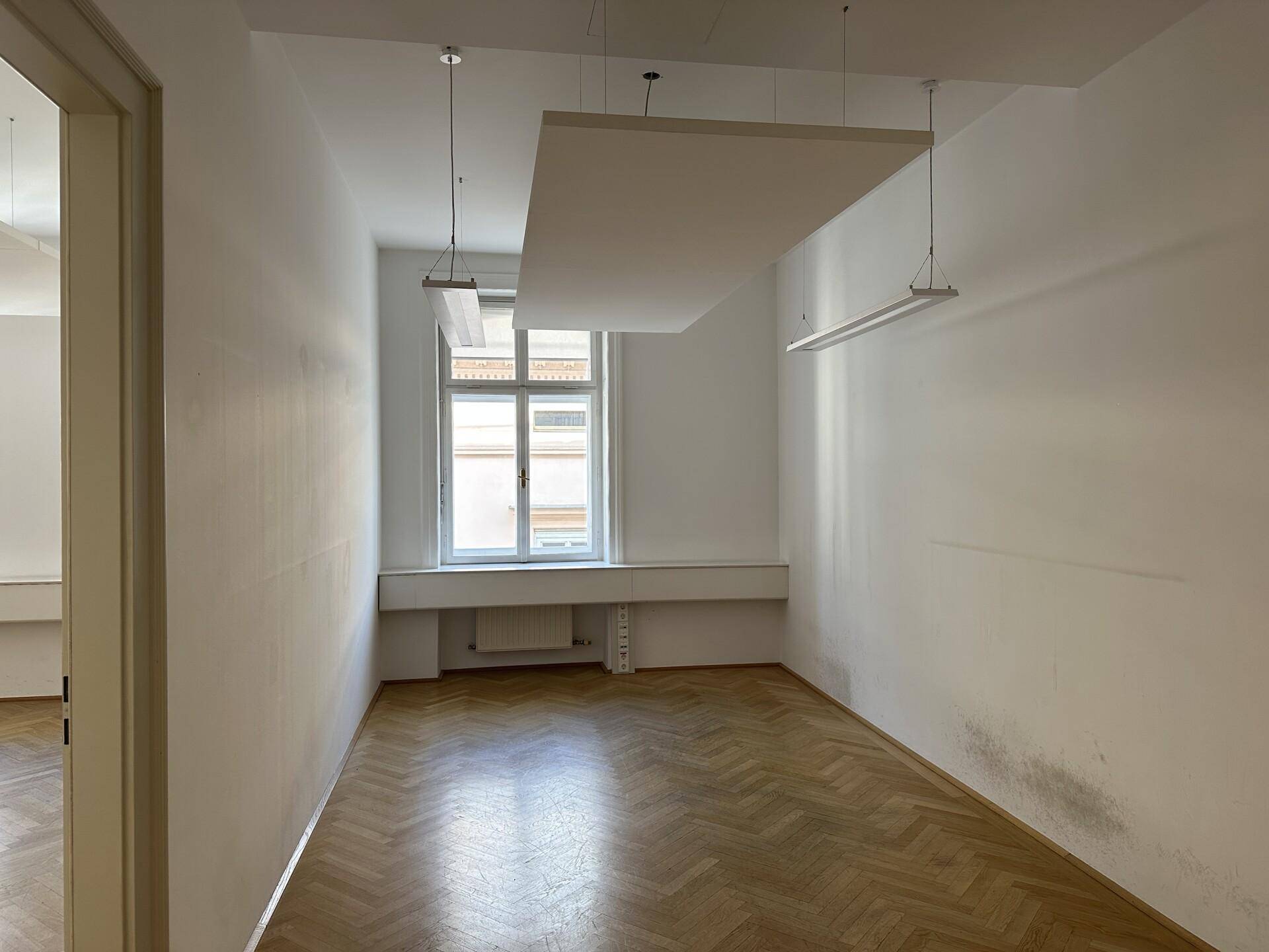 Leeres Büro mit klassischem Fischgrätparkett, einem großen Fenster und modernen Deckenleuchten.