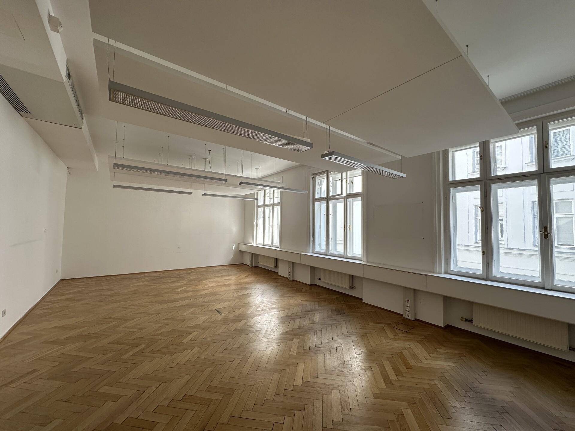 Helles und geräumiges Büro mit Fischgrätparkett, großen Fenstern und modernen Deckenleuchten.