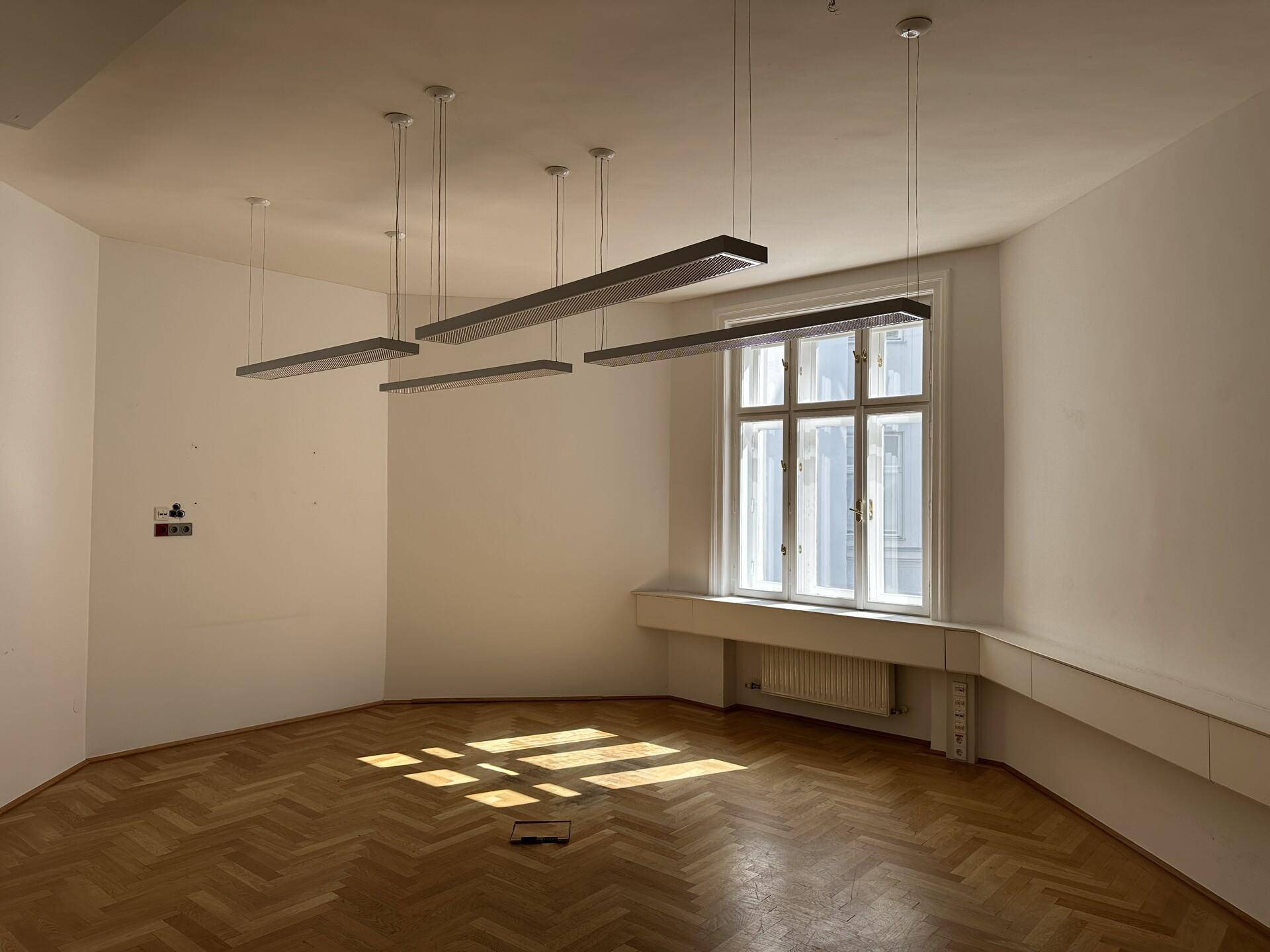 Helles Büro mit Fischgrätparkett, großem Fenster und modernen Hängelampen, durchflutet von Sonnenlicht.
