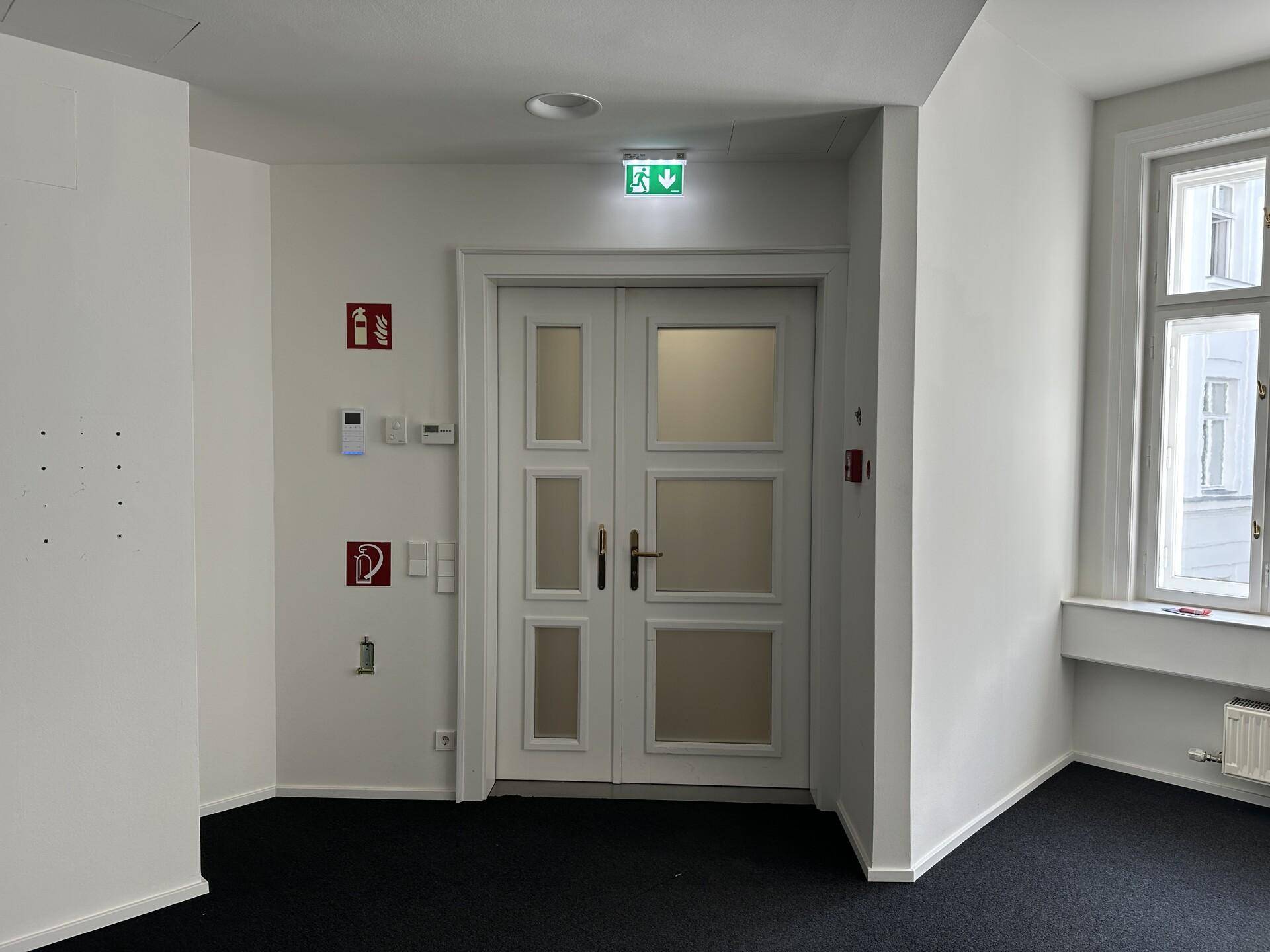 Büroraum mit dunklem Teppichboden, einer Doppeltür und Brandschutzeinrichtungen an der Wand.