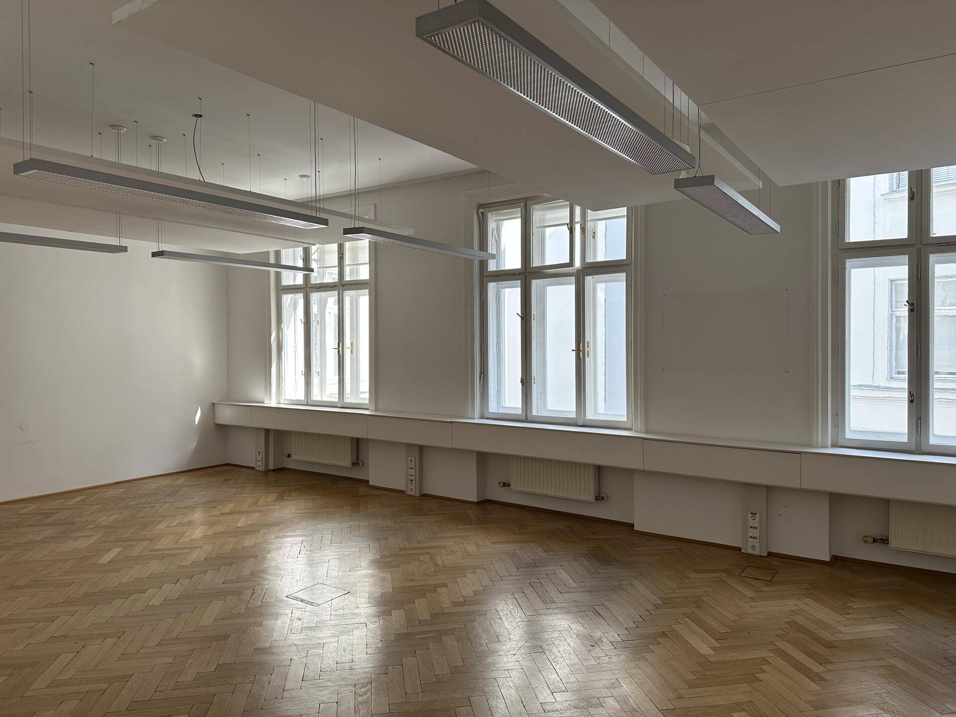 Geräumiges Büro mit Fischgrätparkett, drei großen Fenstern und modernen Hängelampen für eine gute Ausleuchtung.