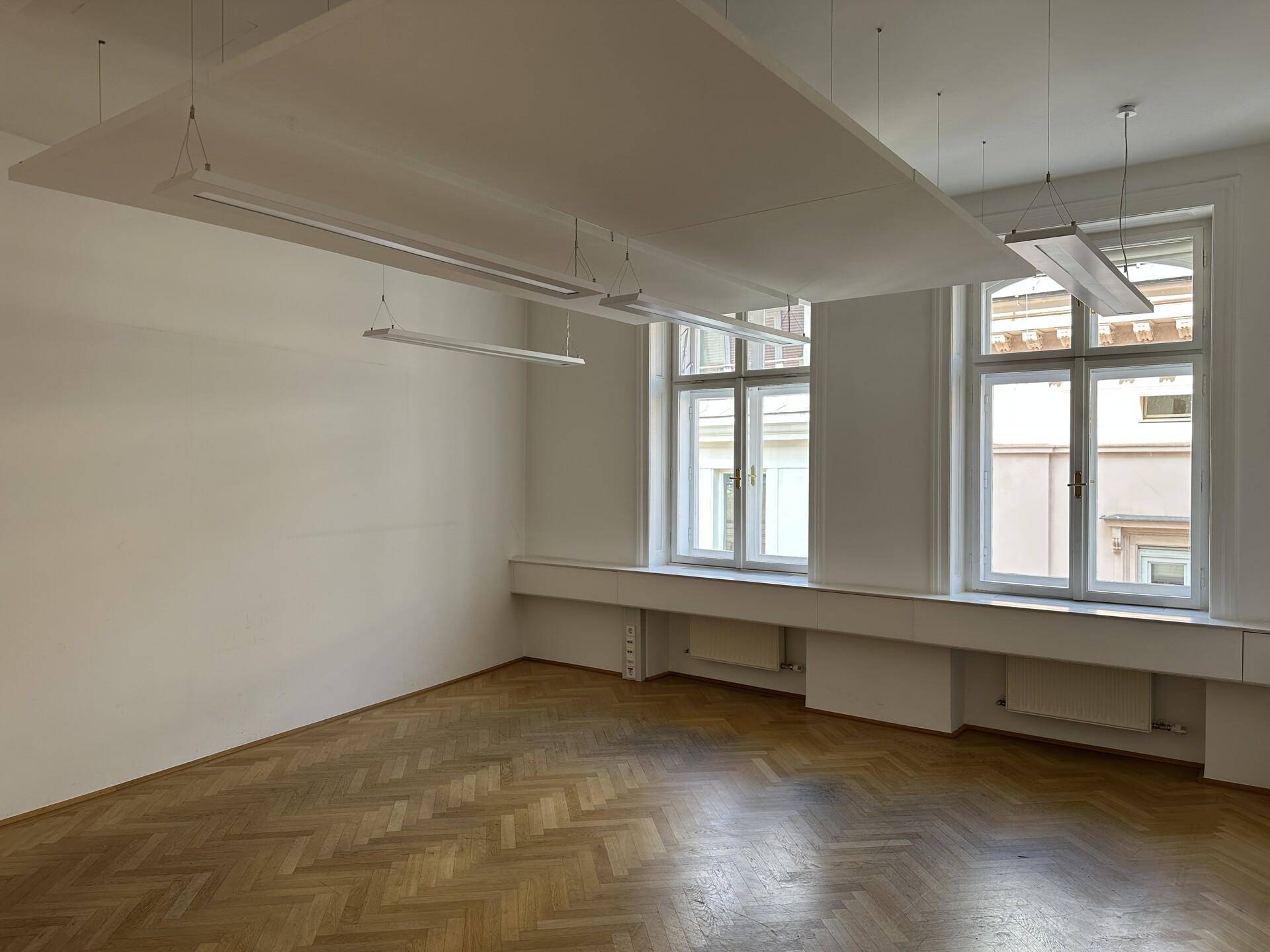 Heller Büroraum mit zwei großen Fenstern, Fischgrätparkett und schwebenden Deckenpaneelen.