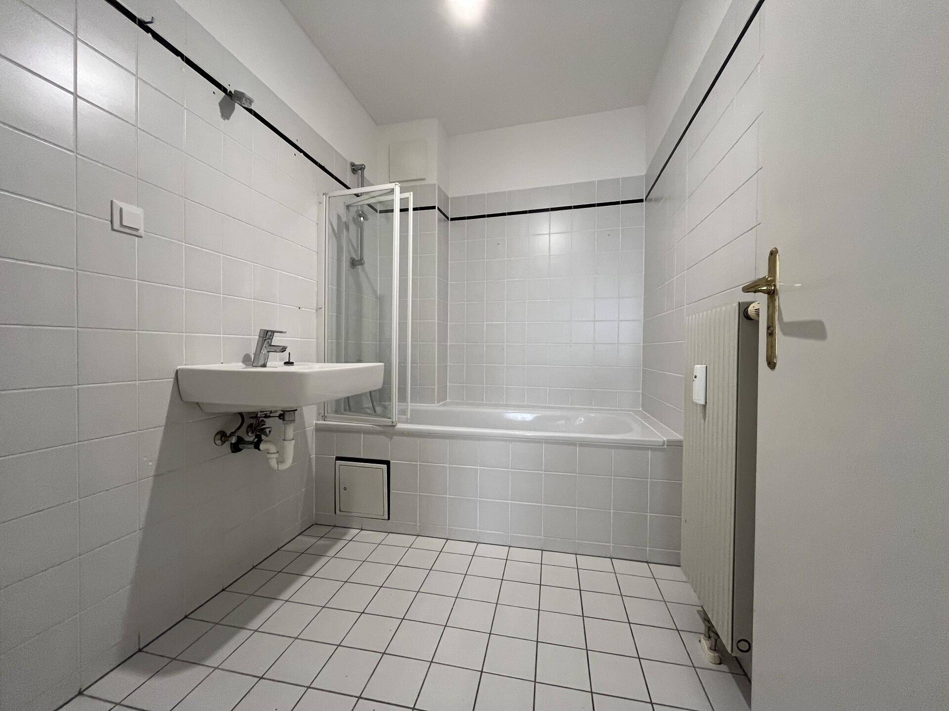 Badezimmer mit weißen Fliesen, Badewanne mit Duschabtrennung und einem einzelnen Waschbecken.