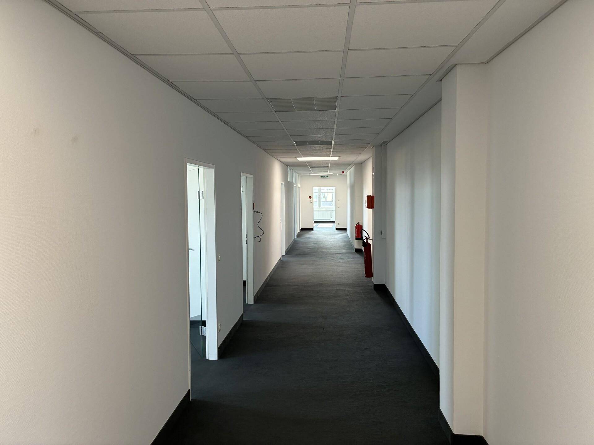 Langer, heller Flur mit weißen Wänden und dunklem Boden, der zu verschiedenen Büroräumen führt.
