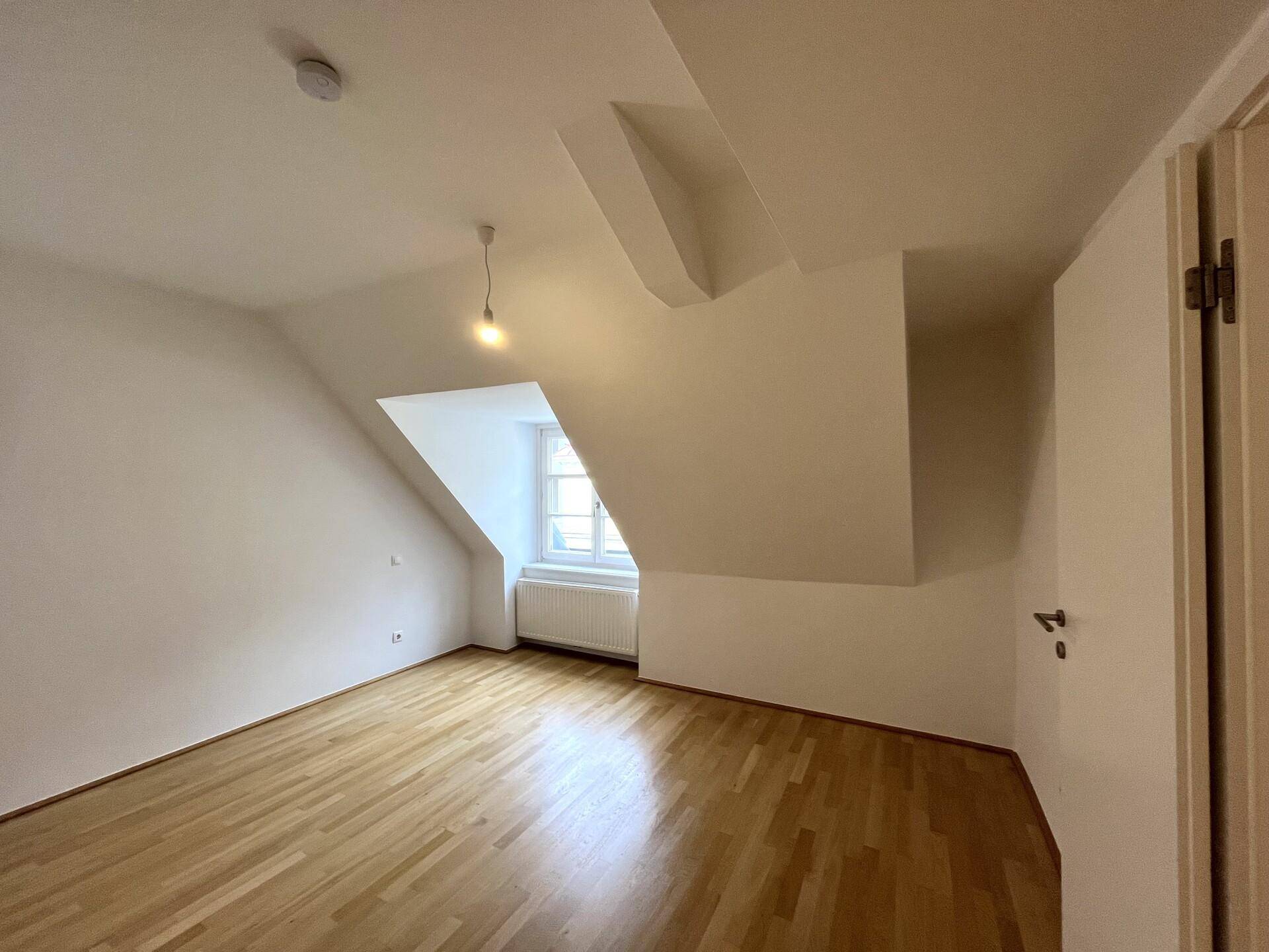 Gemütliches Zimmer im Dachgeschoss mit Parkettboden, schrägen Wänden und einem Fenster, das viel Licht spendet.