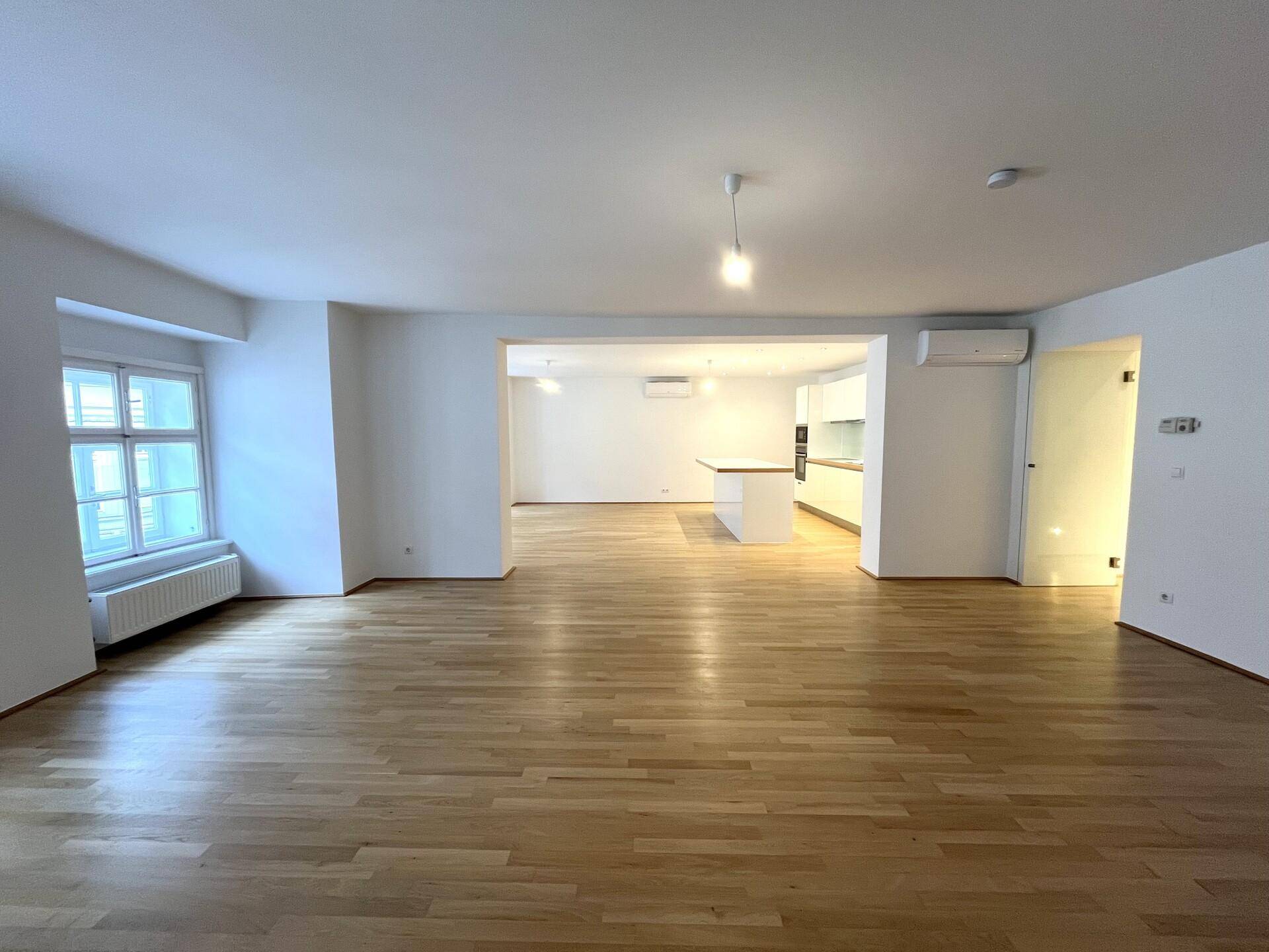 Geräumiger offener Wohn- und Essbereich mit Parkettboden, großen Fenstern und Blick auf die moderne Küche.
