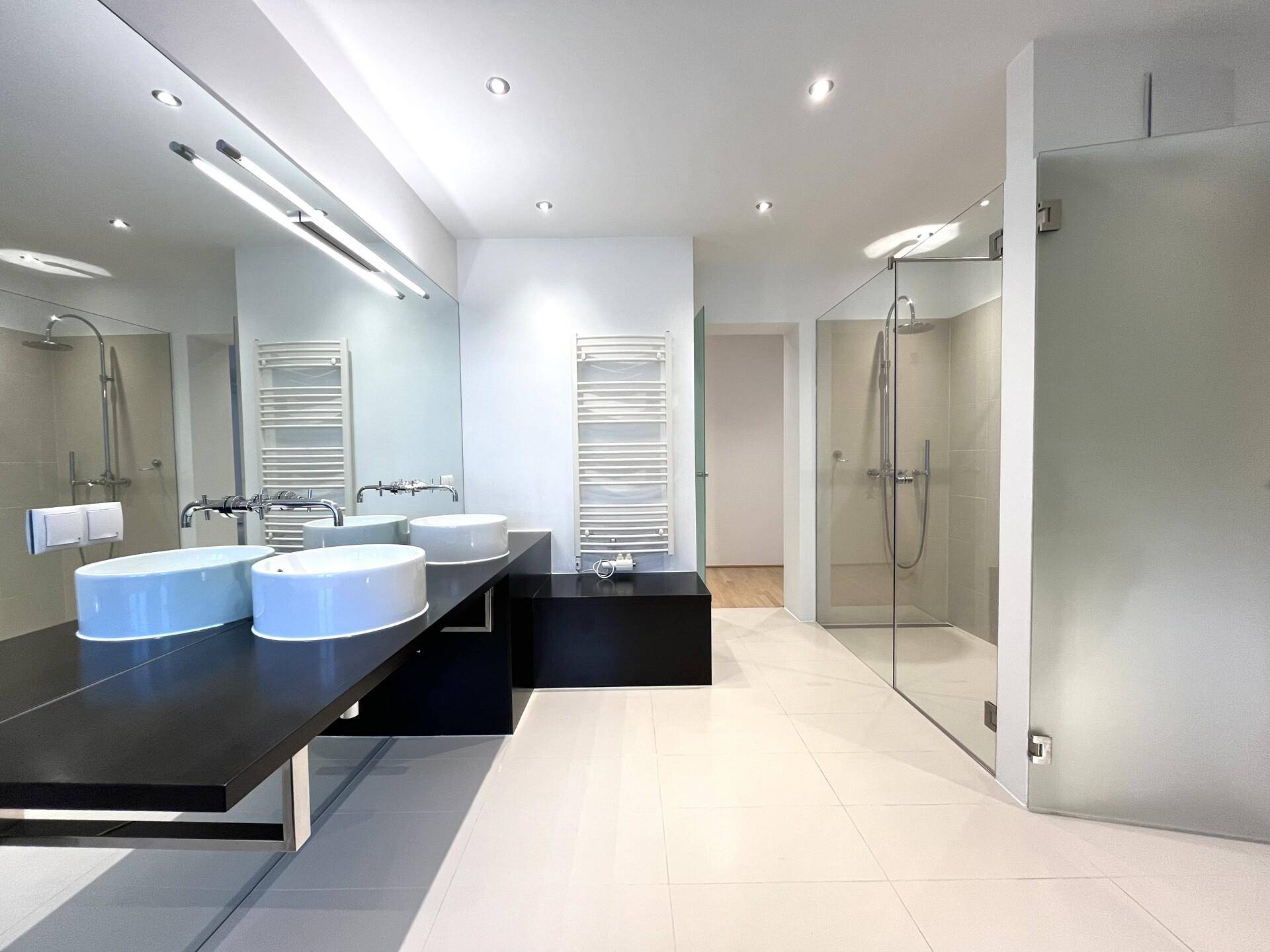 Modernes Badezimmer mit Doppelwaschbecken, großer Spiegelfläche und begehbarer Dusche, hell gefliest.